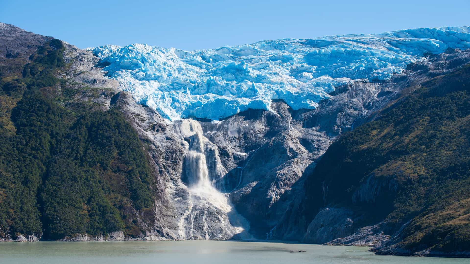 A large glacier on a mountain cliff, with a powerful waterfall cascading down into a fjord in the Chilean Fjords.