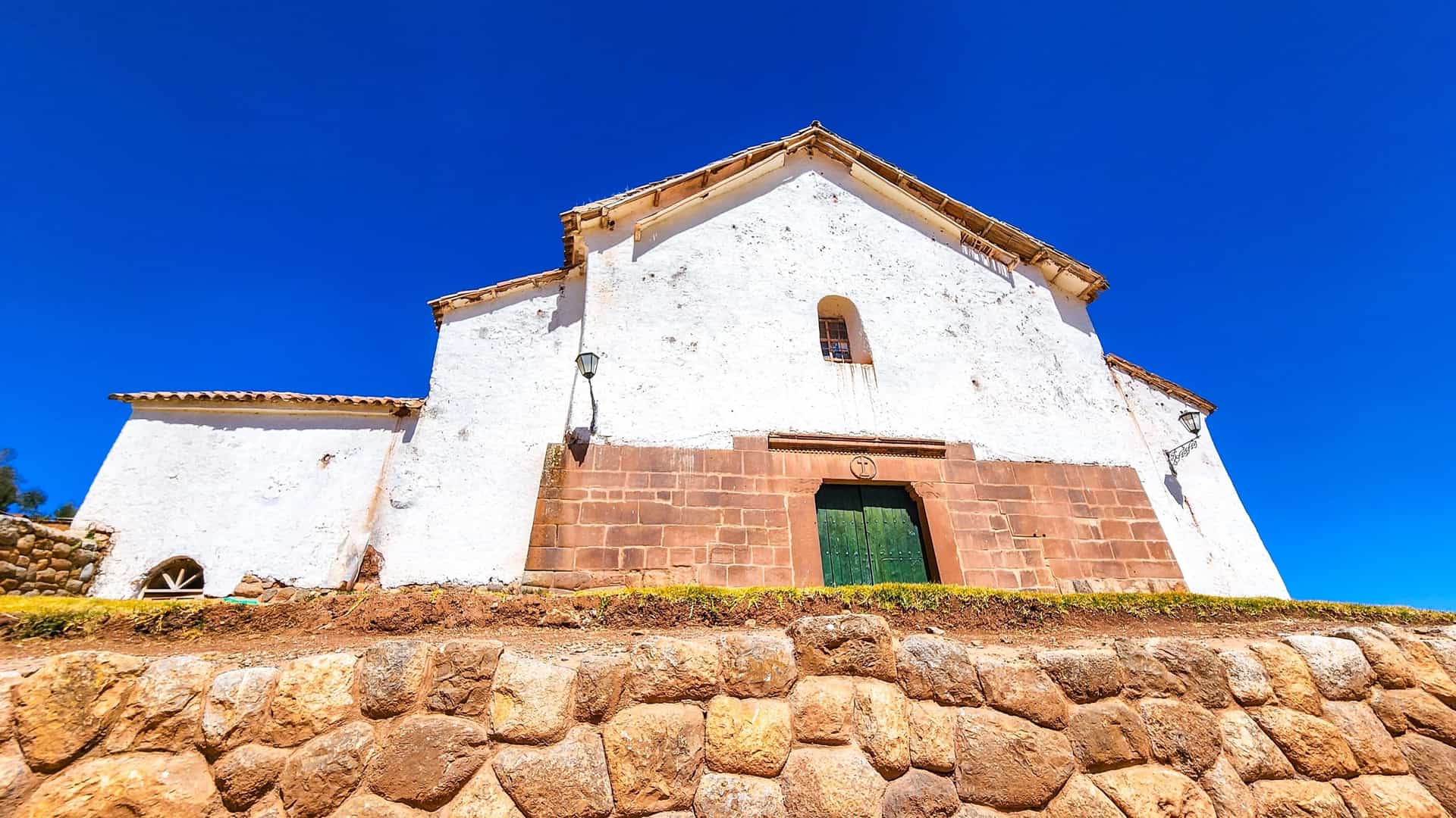 A low-angle view shows the facade of a church or colonial building in Chinchero, Peru, with a white upper section and a base of reddish-brown Inca stone blocks, all sitting atop a large, rough stone retaining wall under a brilliant, clear blue sky.