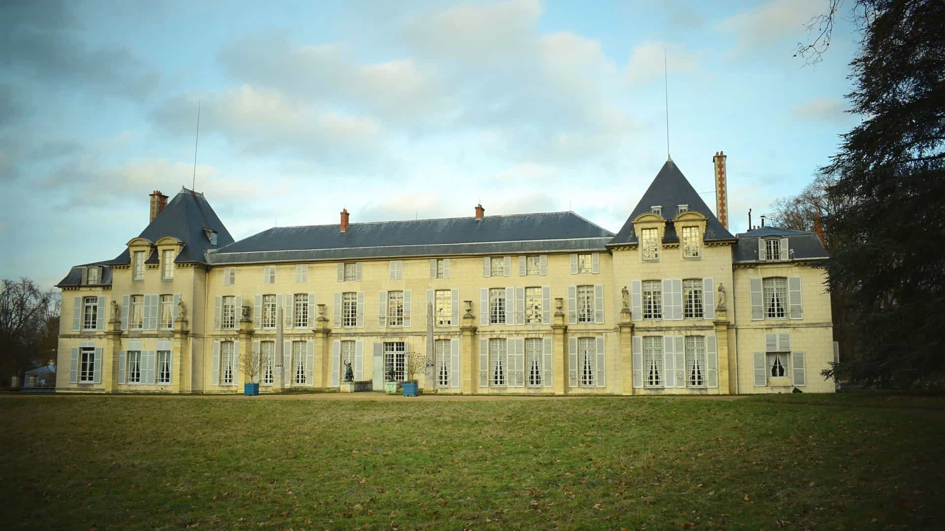 A long, horizontal shot of the Chateau de Malmaison's classicist facade with a gray roof and white shutters, surrounded by a green lawn in France.