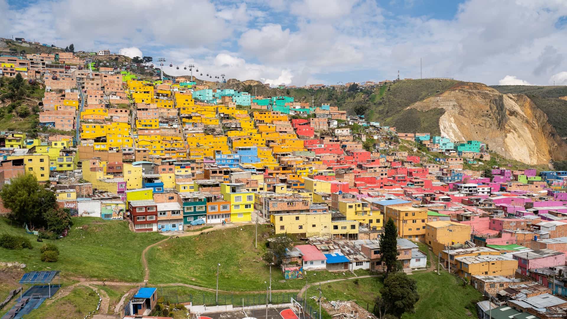 A colorful residential area of Ciudad Bolivar, Bogotá, with bright yellow, blue, and pink houses built on a hillside, with a cable car visible in the sky.