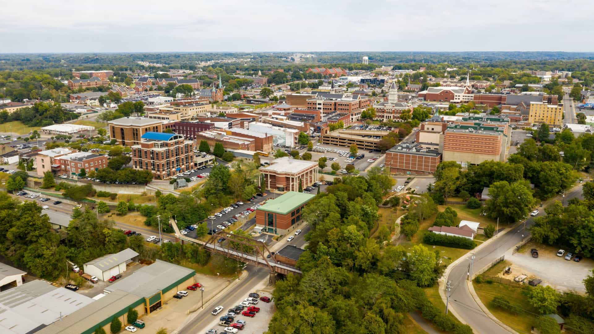 An aerial view of Clarksville, Tennessee, showing its downtown area with buildings, streets, and parking lots surrounded by green trees.