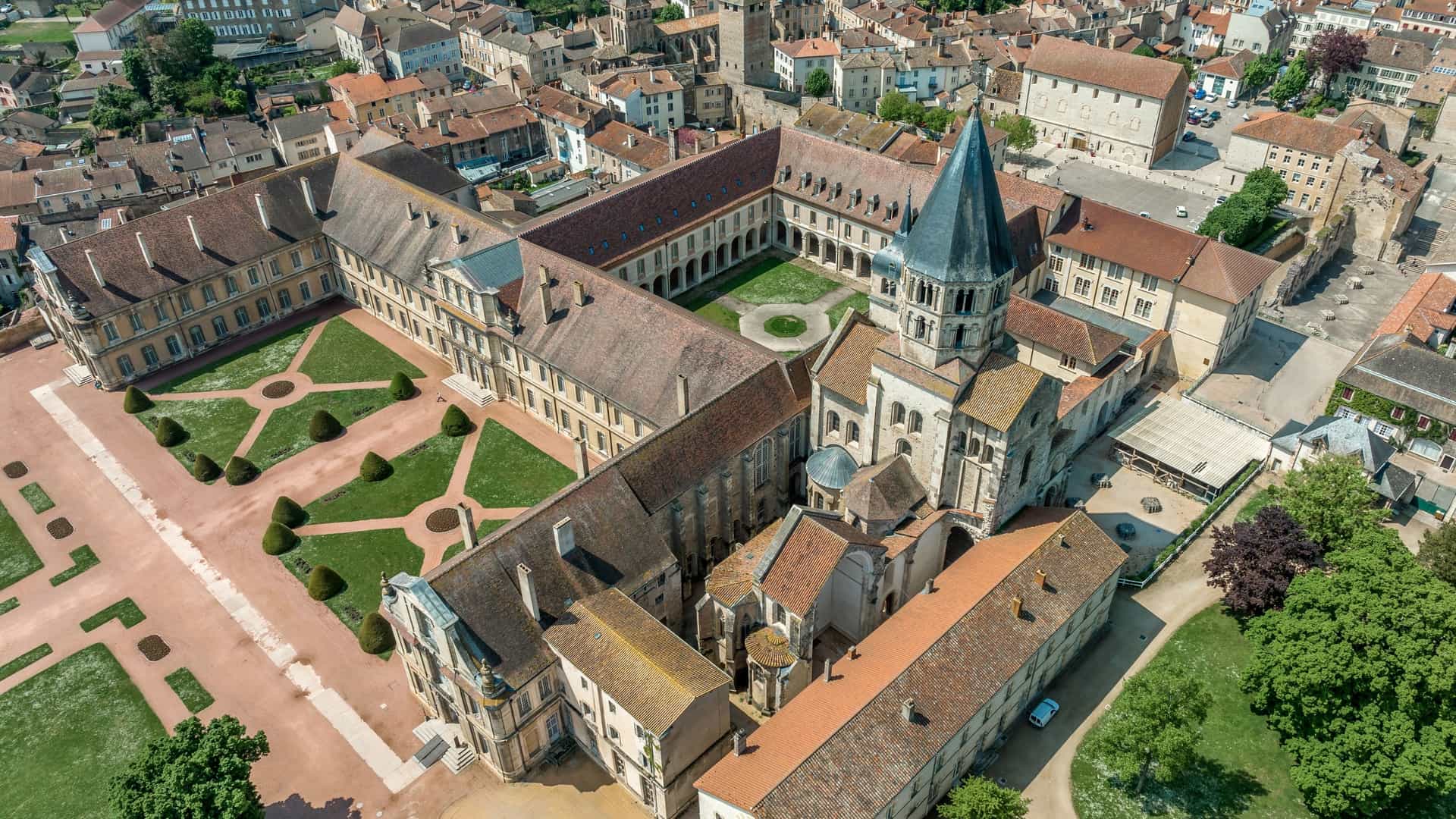 An aerial view shows the extensive architectural complex of Cluny Abbey, France, with the abbey ruins and surrounding buildings in a dense townscape.