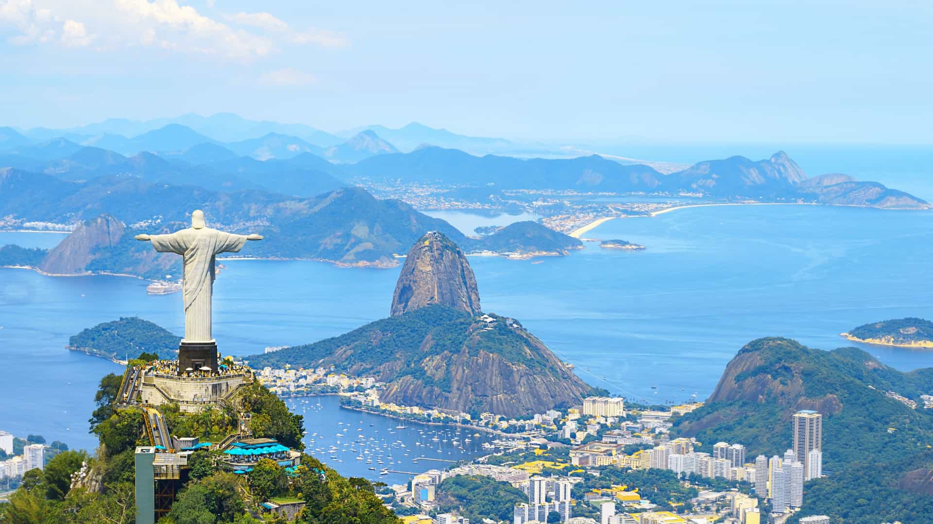 A stunning panoramic view of Rio de Janeiro's coastline, with the iconic Christ the Redeemer statue in the foreground, Sugarloaf Mountain, and the sprawling city.