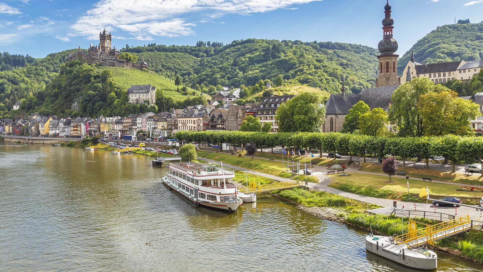 A beautiful view of Cochem, Germany, showing the historic Reichsburg Castle on a hill, the town's colorful buildings, and a cruise boat on the Moselle River.