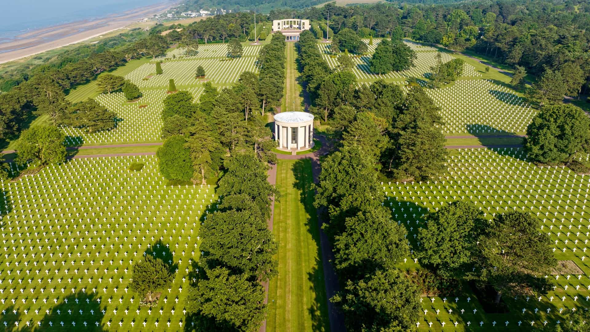 An aerial view of the Normandy American Cemetery and Memorial in Colleville-sur-Mer, showing rows of white crosses and stars of David extending across the green landscape toward the ocean.