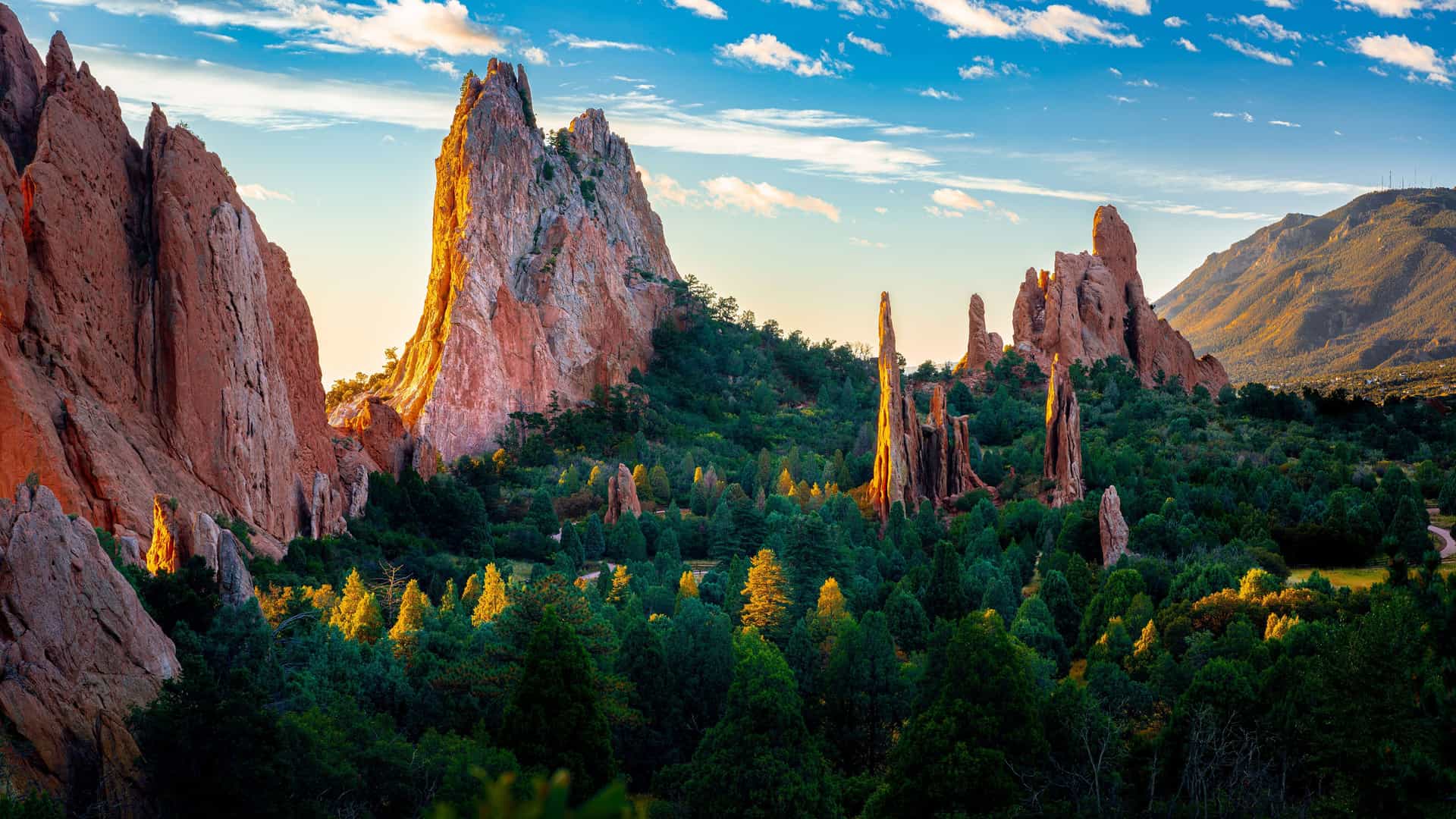 "A scenic landscape of dramatic red rock formations and towering sandstone spires surrounded by vibrant green trees under a bright blue sky at the Garden of the Gods Park in Colorado Springs.  "