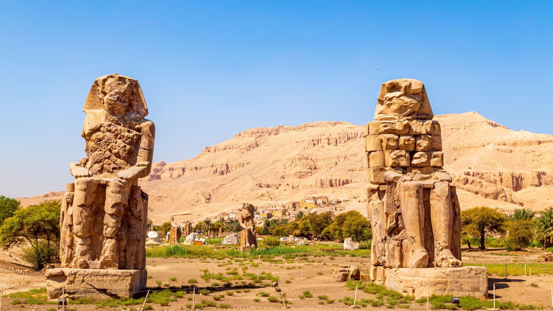 Two colossal statues of Amenhotep III, known as the Colossi of Memnon, stand prominently in a desert landscape with hills and green trees in the background.