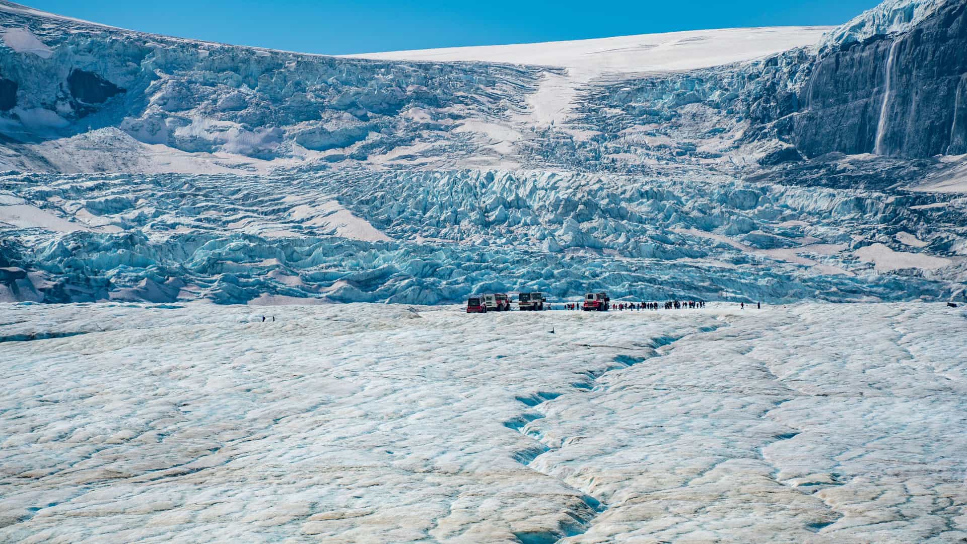 "A scenic shot of the vast Columbia Icefield in Jasper National Park, Canada, with a line of red Ice Explorer tour buses and tourists on the surface of the stunning Athabasca Glacier. "