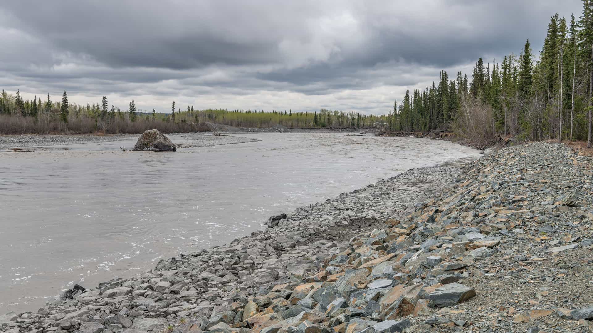 "A view of the wide, gray-colored Copper River in Alaska, with a rocky shore and evergreen trees lining the banks under a dramatic, cloudy sky. "