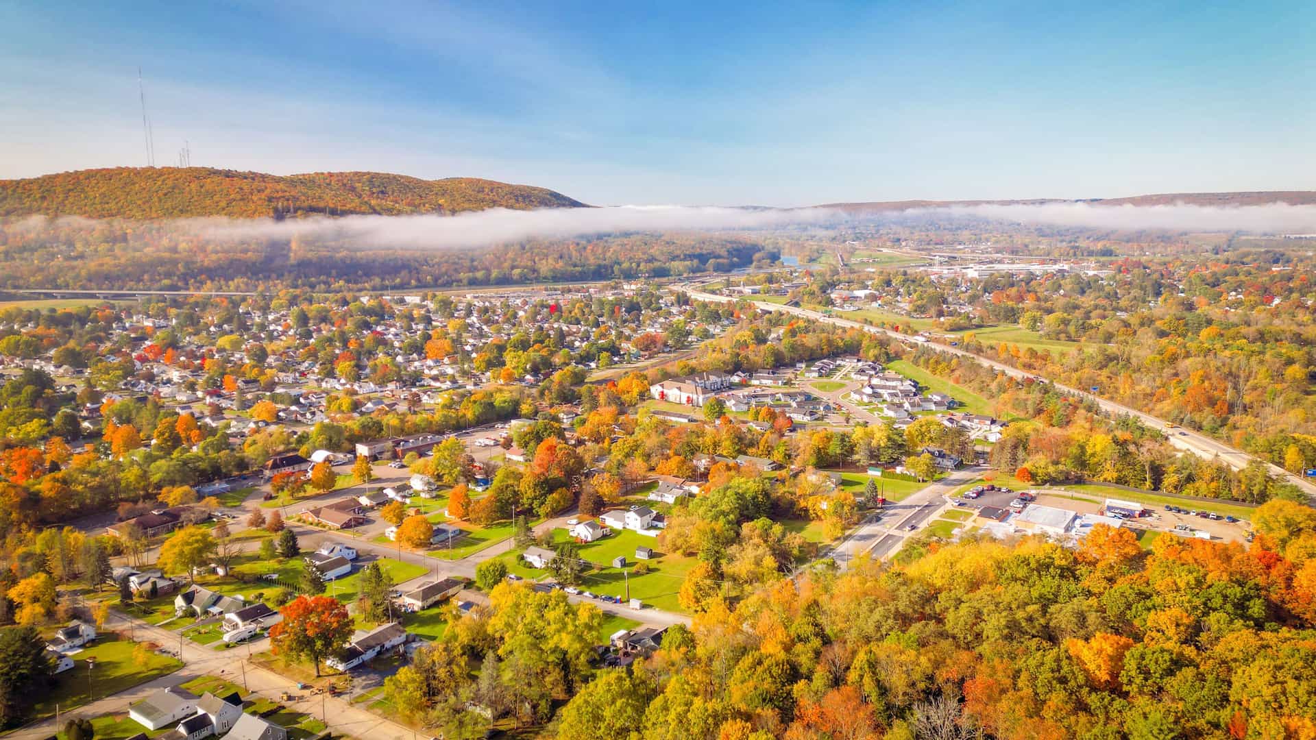 "An aerial view of the town of Corning, New York, in the autumn, with houses and streets surrounded by a vibrant tapestry of orange and yellow foliage, and low-lying clouds hanging over the forested hills in the distance.  "