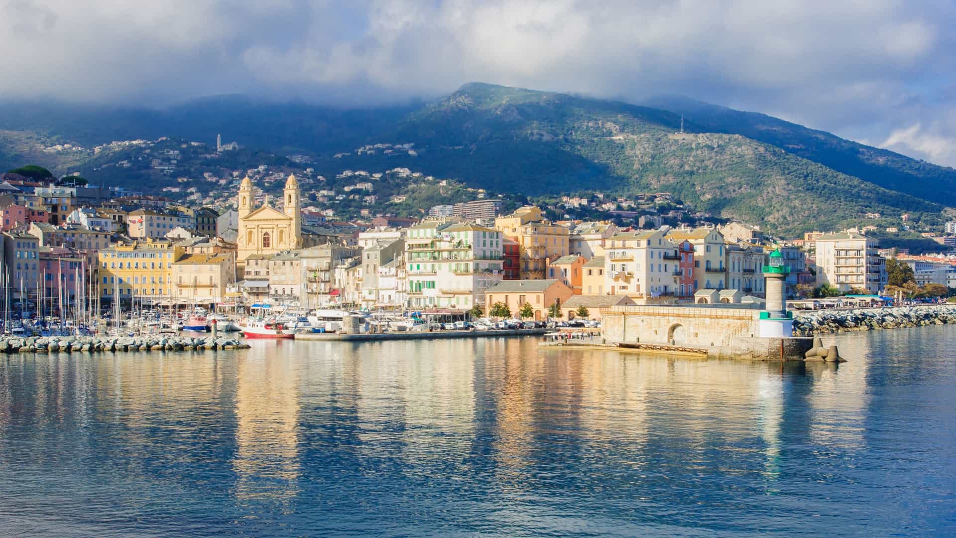 A picturesque view of a coastal city in Corsica, with a bustling harbor, a green lighthouse, and colorful buildings at the foot of a large mountain.