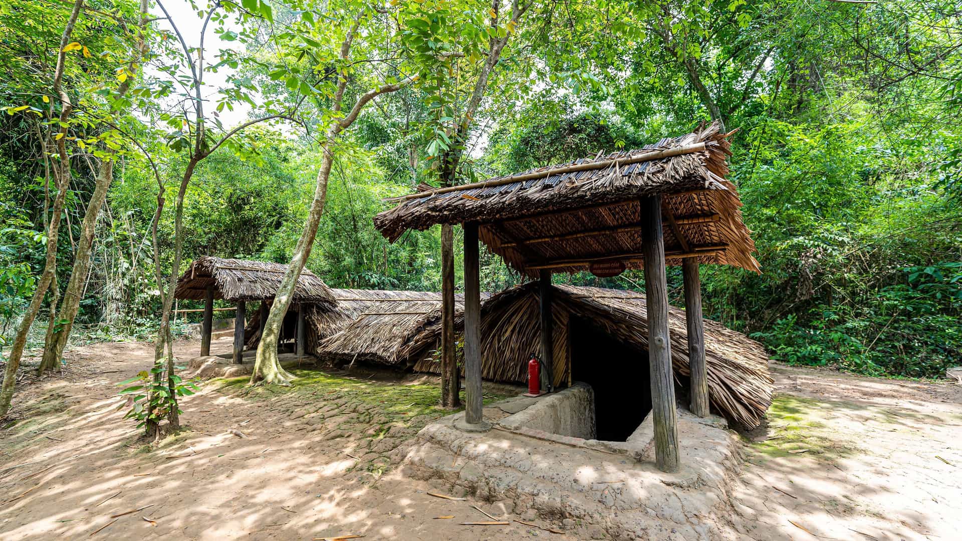 A small, camouflaged entrance to the Cu Chi Tunnels, a historic military site, is covered by a thatched hut in a dense, green jungle in Vietnam.