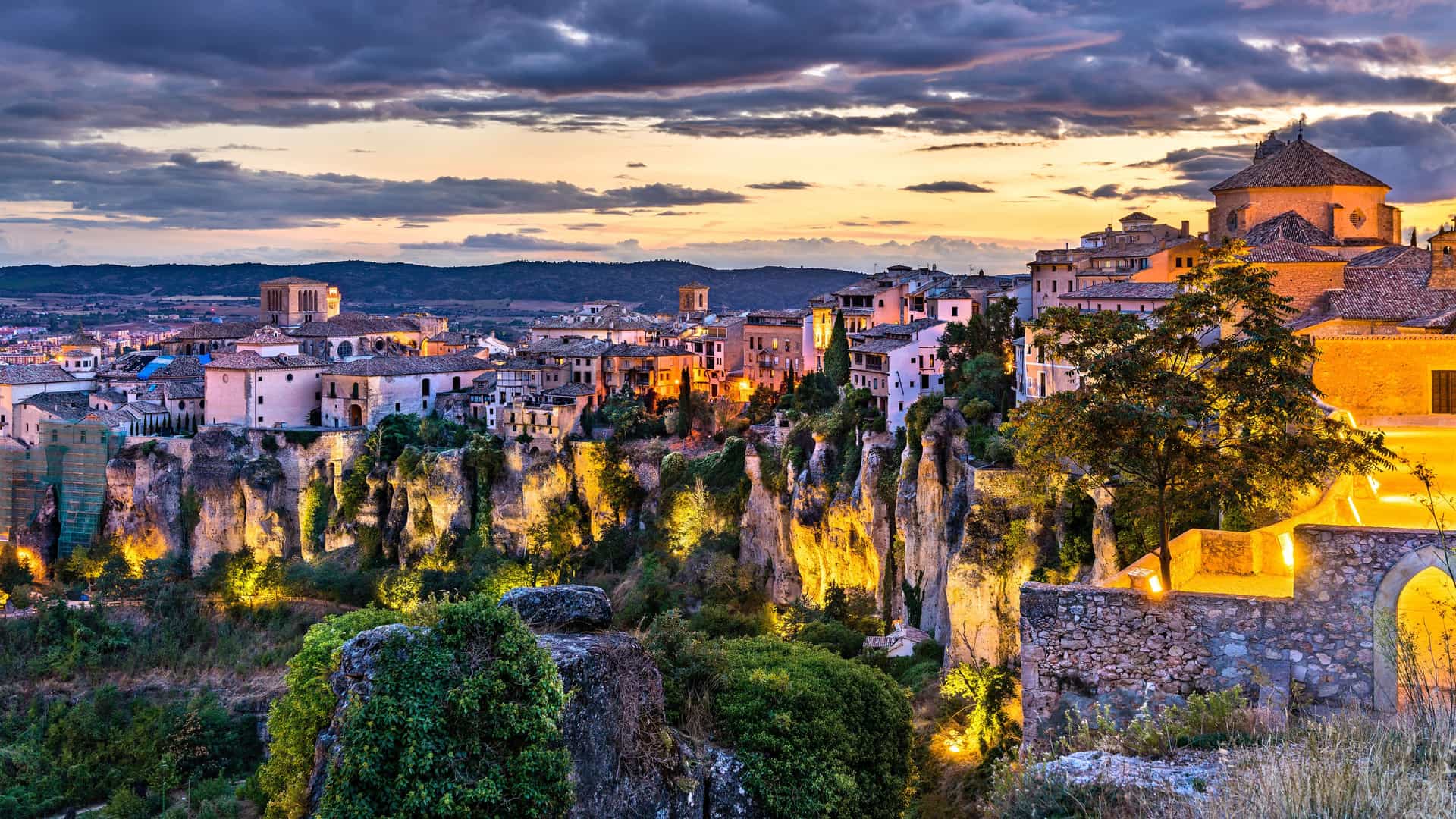 "A breathtaking image of the historic city of Cuenca, Spain, at dusk, showing the famous ""Hanging Houses"" built precariously on the cliffs, illuminated against the dramatic sunset sky.  "