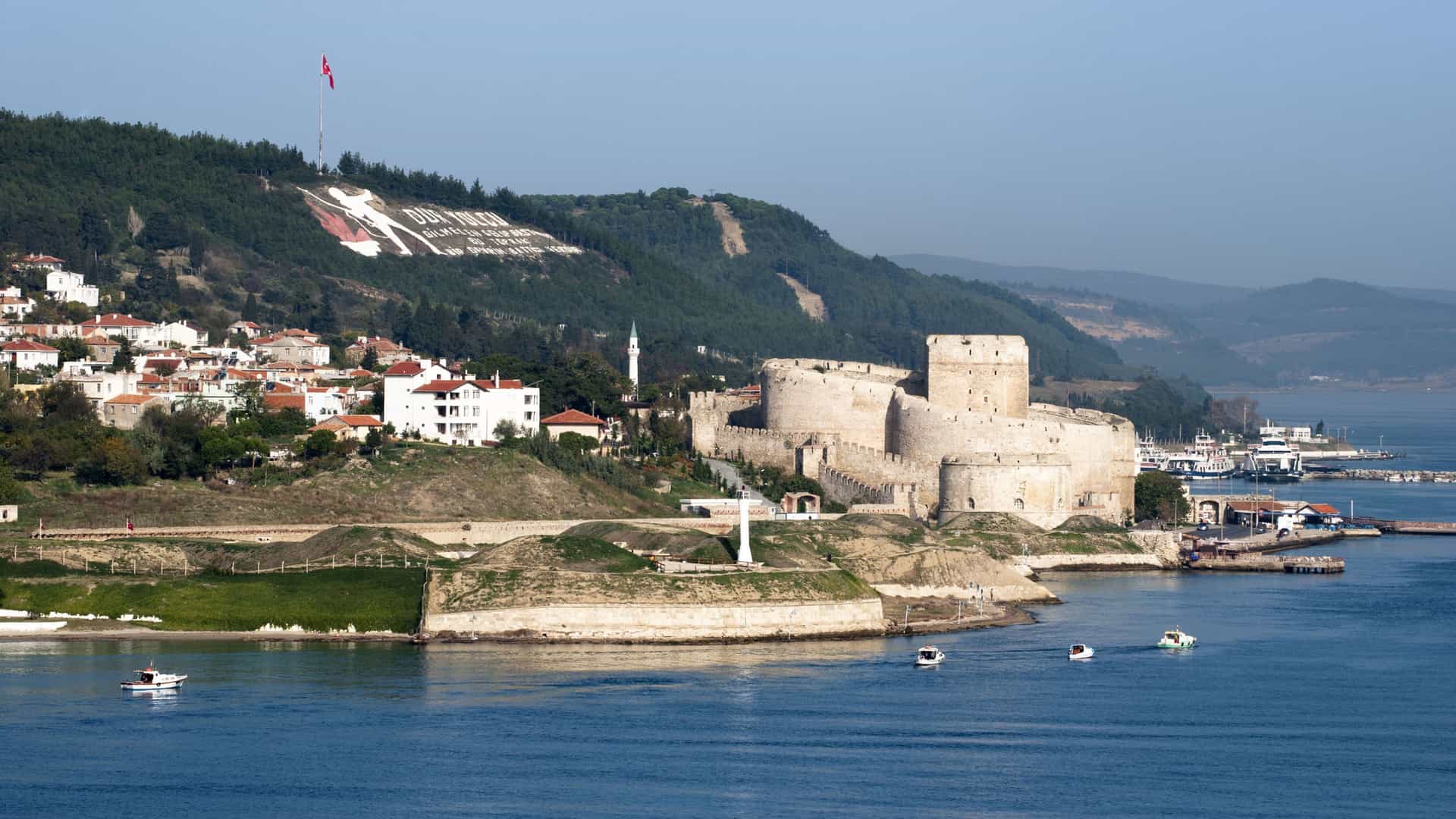A scenic view of the historic Kilitbahir Castle fortress on the Dardanelles Strait in Turkey, situated on a hillside above a harbor and coastal town.