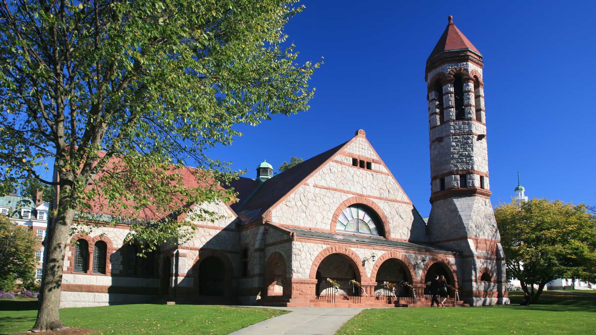 A wide-angle shot of the historic Rollins Chapel at Dartmouth College, a stone building with a prominent bell tower and red roof on a sunny day.