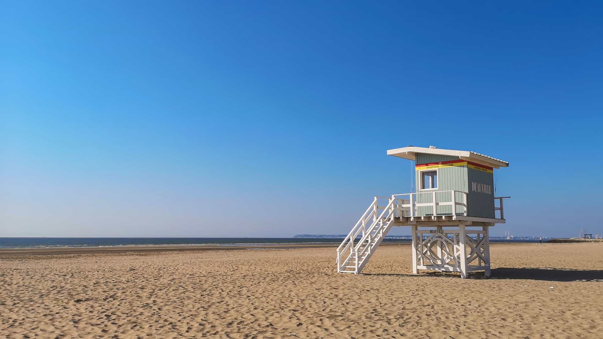 "A small, light green and white lifeguard hut with a wooden staircase sits on the wide, sandy beach of Deauville, France, with a calm blue sea and a clear sky stretching to the horizon.  "