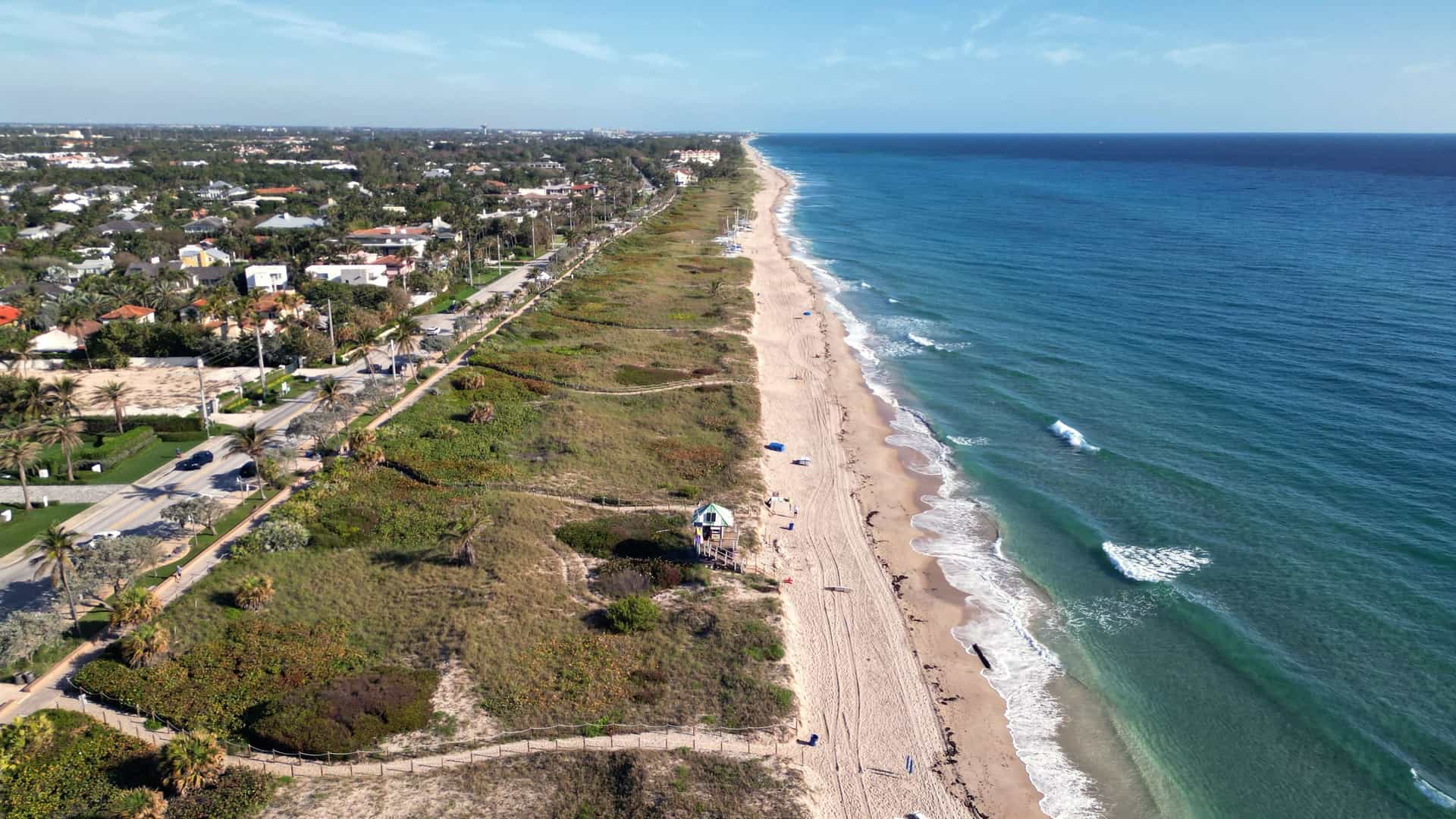 "An aerial view of the beautiful sandy beach and turquoise ocean of Delray Beach, Florida, with a lifeguard stand in the middle ground and a line of palm trees and coastal homes on the shoreline.  "