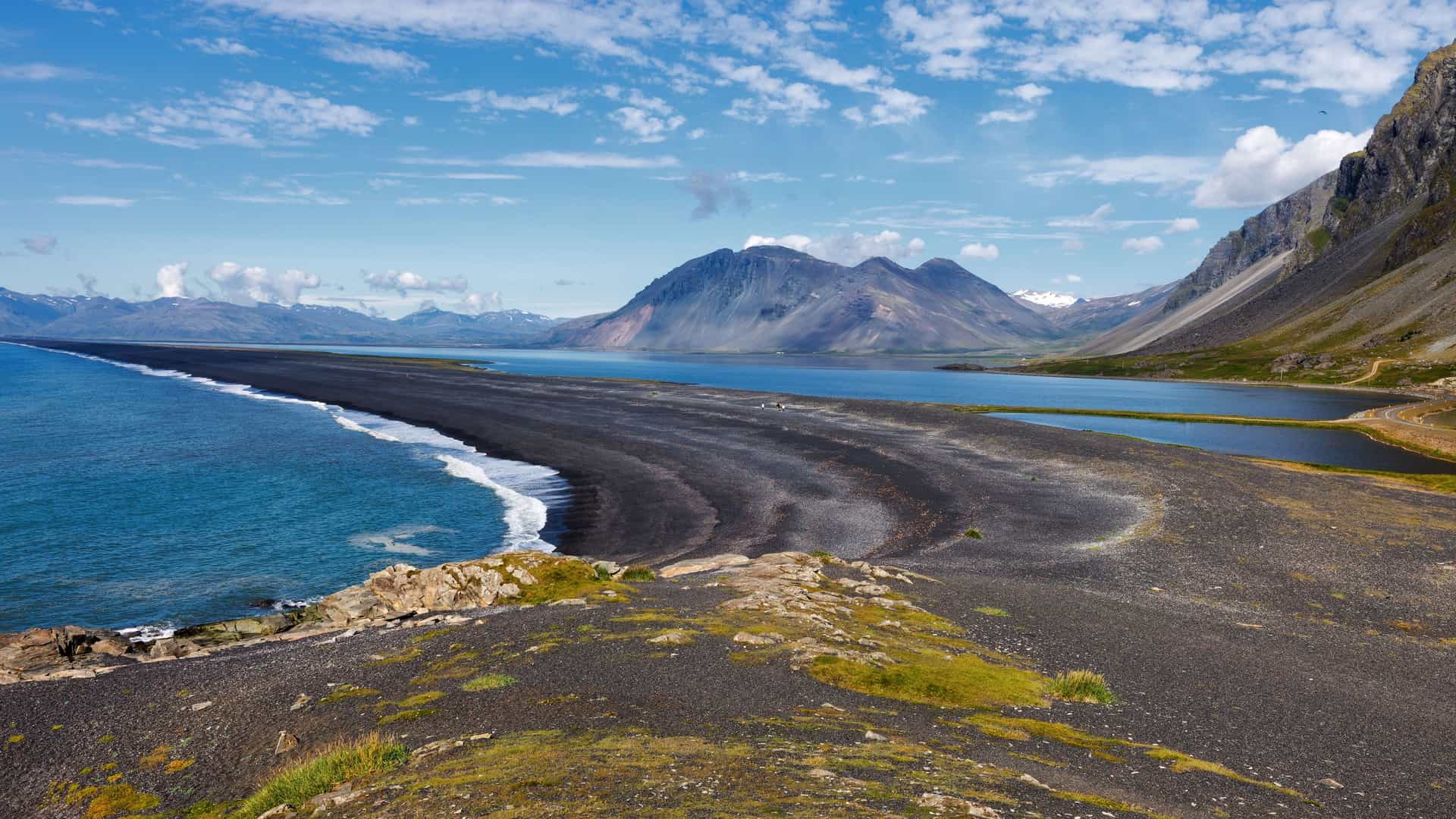 A panoramic view of the dramatic black sand beach and rugged coastline near Djúpivogur, Iceland, with a sweeping mountain range in the background.