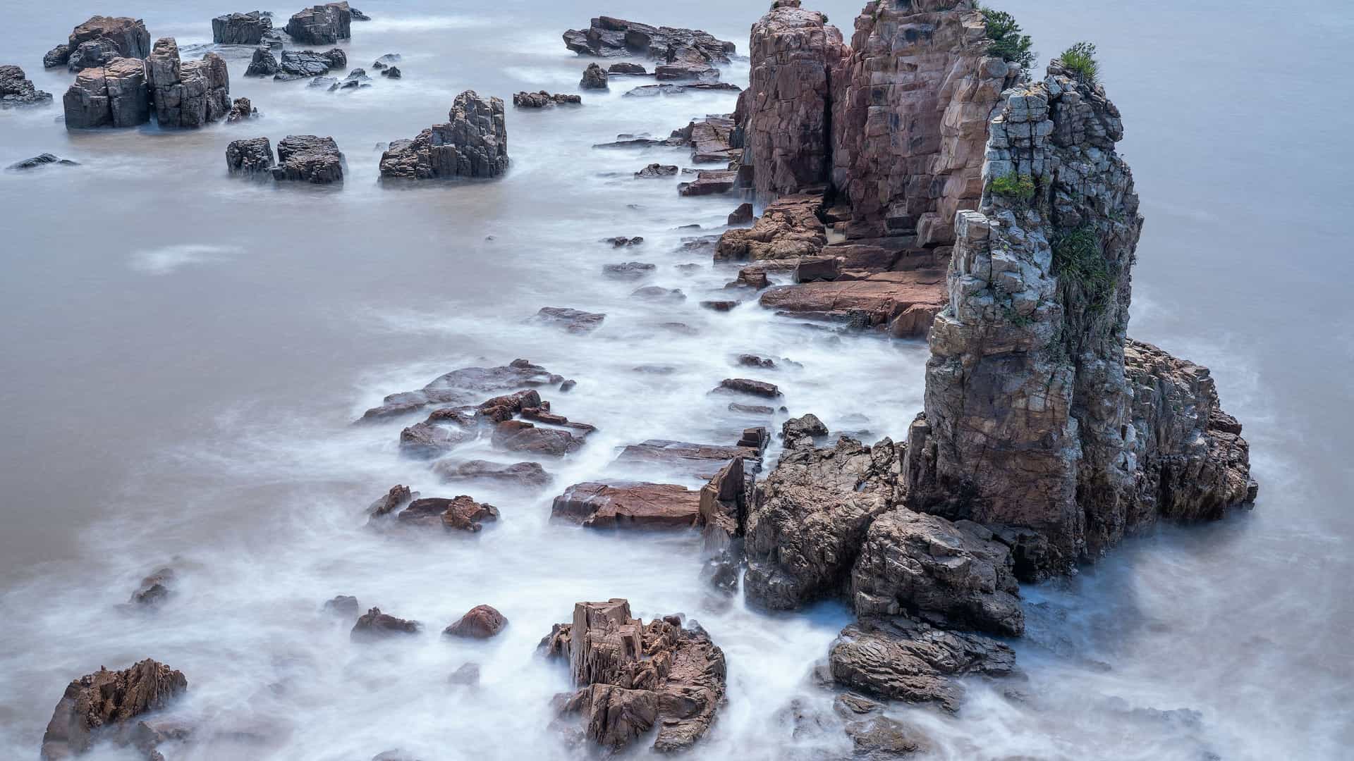 The dramatic, rocky coastline of Dongtou, China, with jagged rock formations and sea stacks surrounded by the misty, blurred effect of a long-exposure photograph.