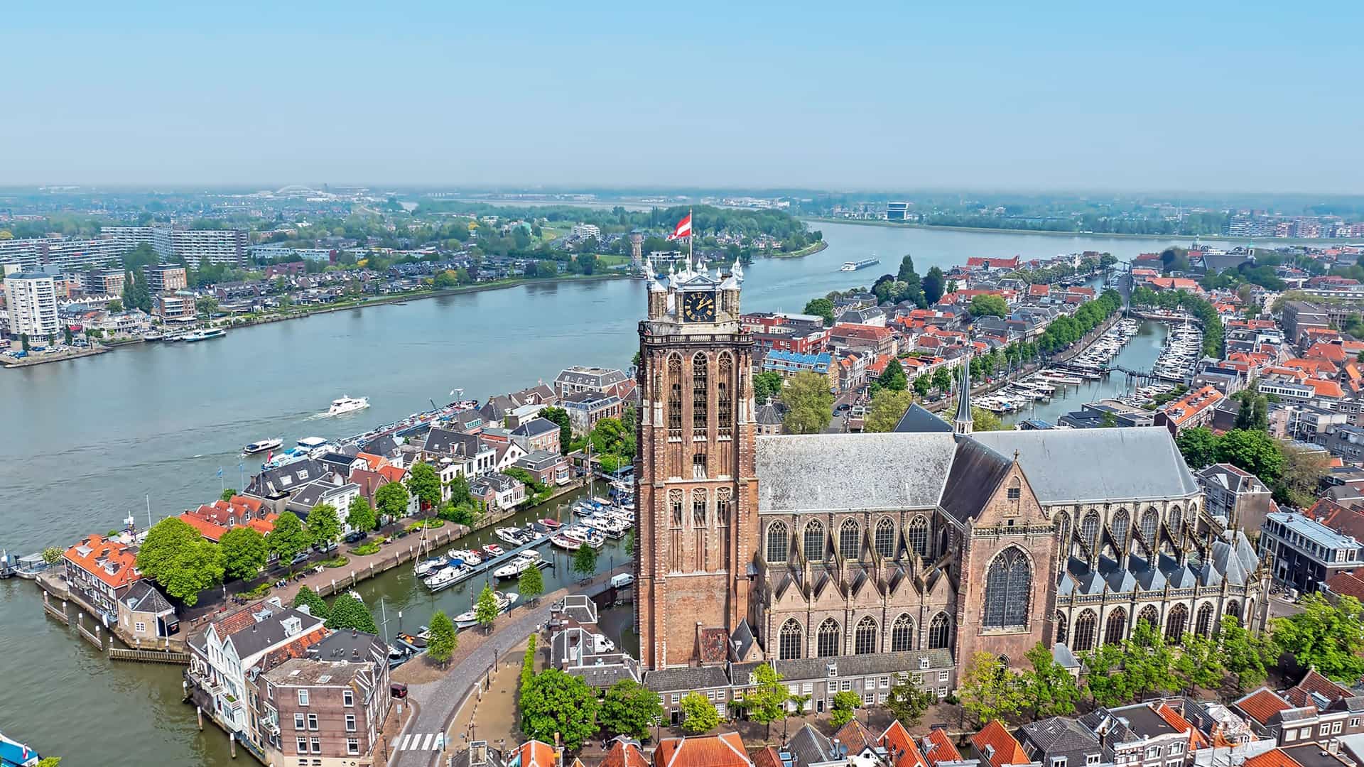 An aerial view of the historic Dutch city of Dordrecht, showcasing the iconic Grote Kerk and its bell tower overlooking a bustling network of rivers and canals.