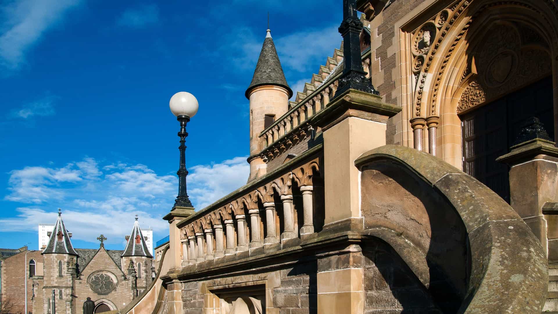 A historic stone building in Dundee, Scotland, with a large ornate archway, a turret, and a lamp post, set against a bright blue sky with white clouds.