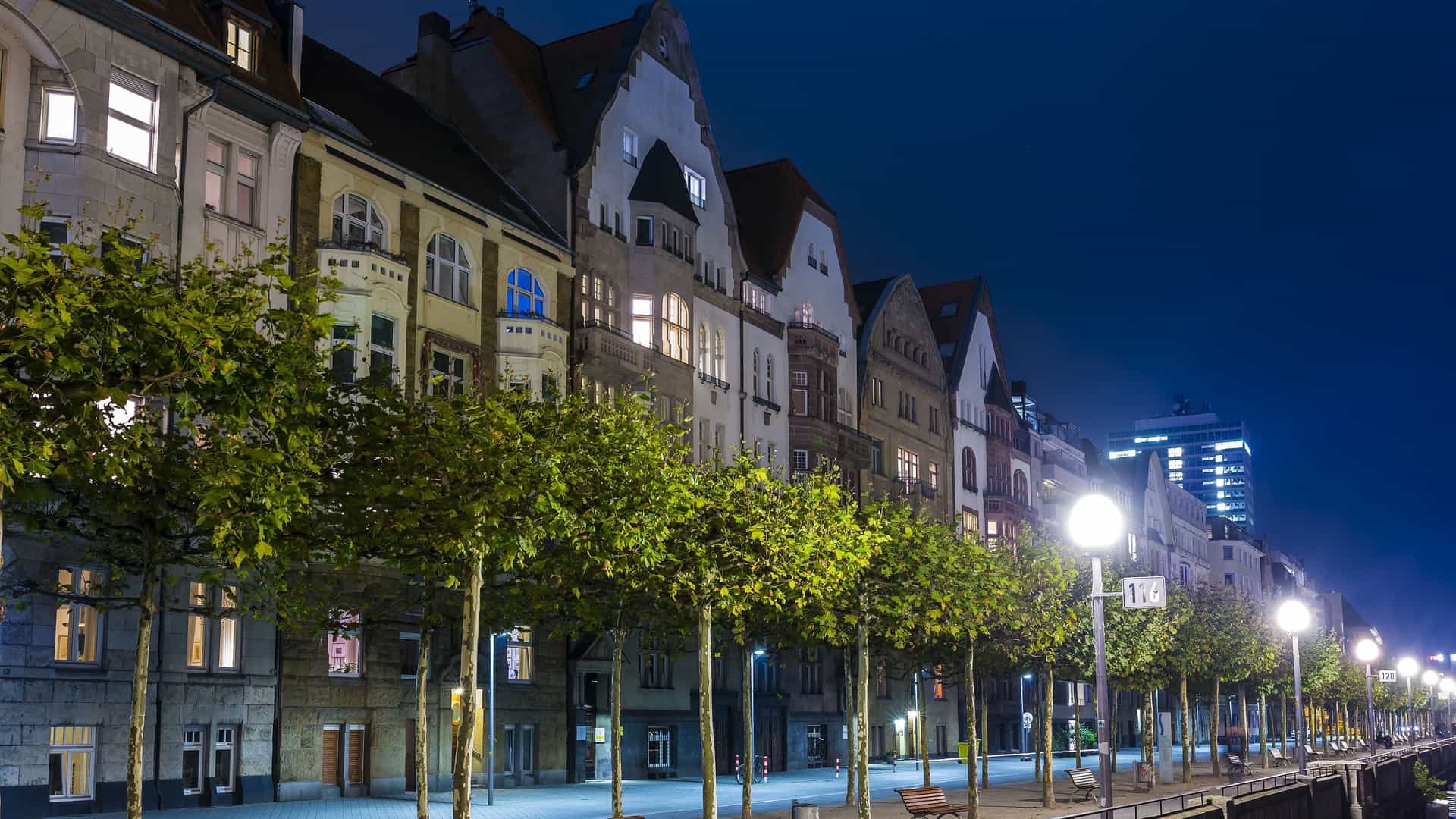 A serene nighttime shot of a street in Düsseldorf, Germany, with a line of trees and old buildings with glowing windows lining the street under a deep blue sky.