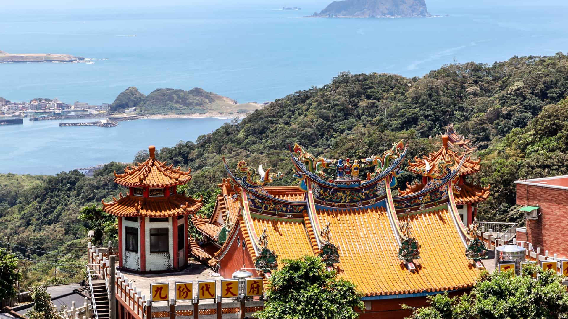 A high-angle view of a traditional Chinese temple with ornate orange and green roofs, overlooking the vast East China Sea with forested mountains and islands in the distance.