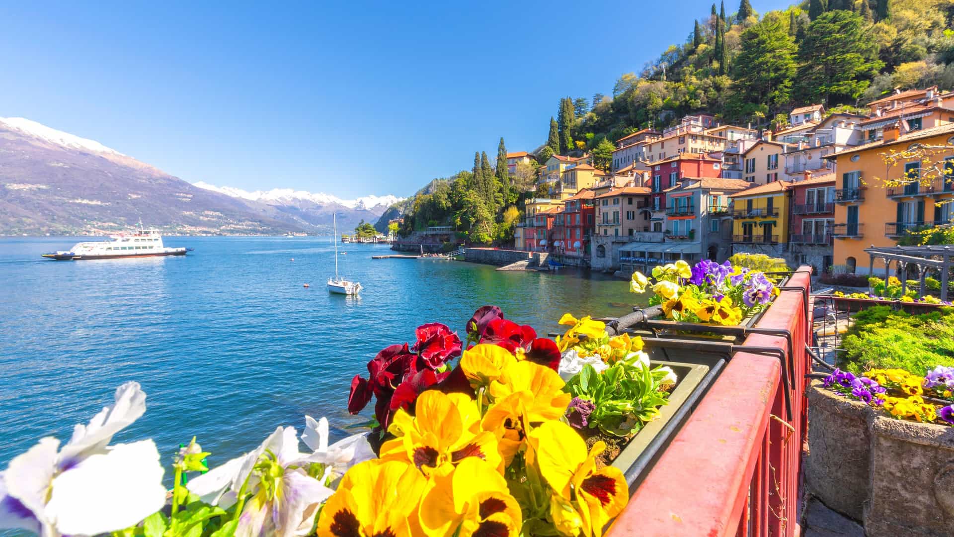 A scenic view of a lakeshore town in Eaux-Rousses, Italy, with colorful buildings nestled on a hillside above the deep blue lake, with snow-capped mountains in the background.