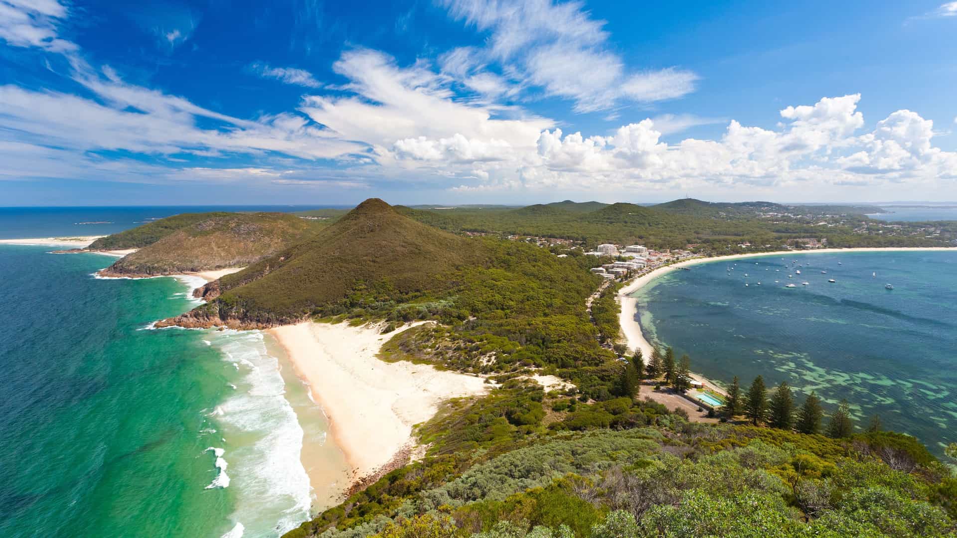 A high-angle panoramic shot of the beautiful coastline near Eden, Australia, with a pristine sandy beach, rugged hills, and a tranquil bay dotted with boats under a bright blue sky.