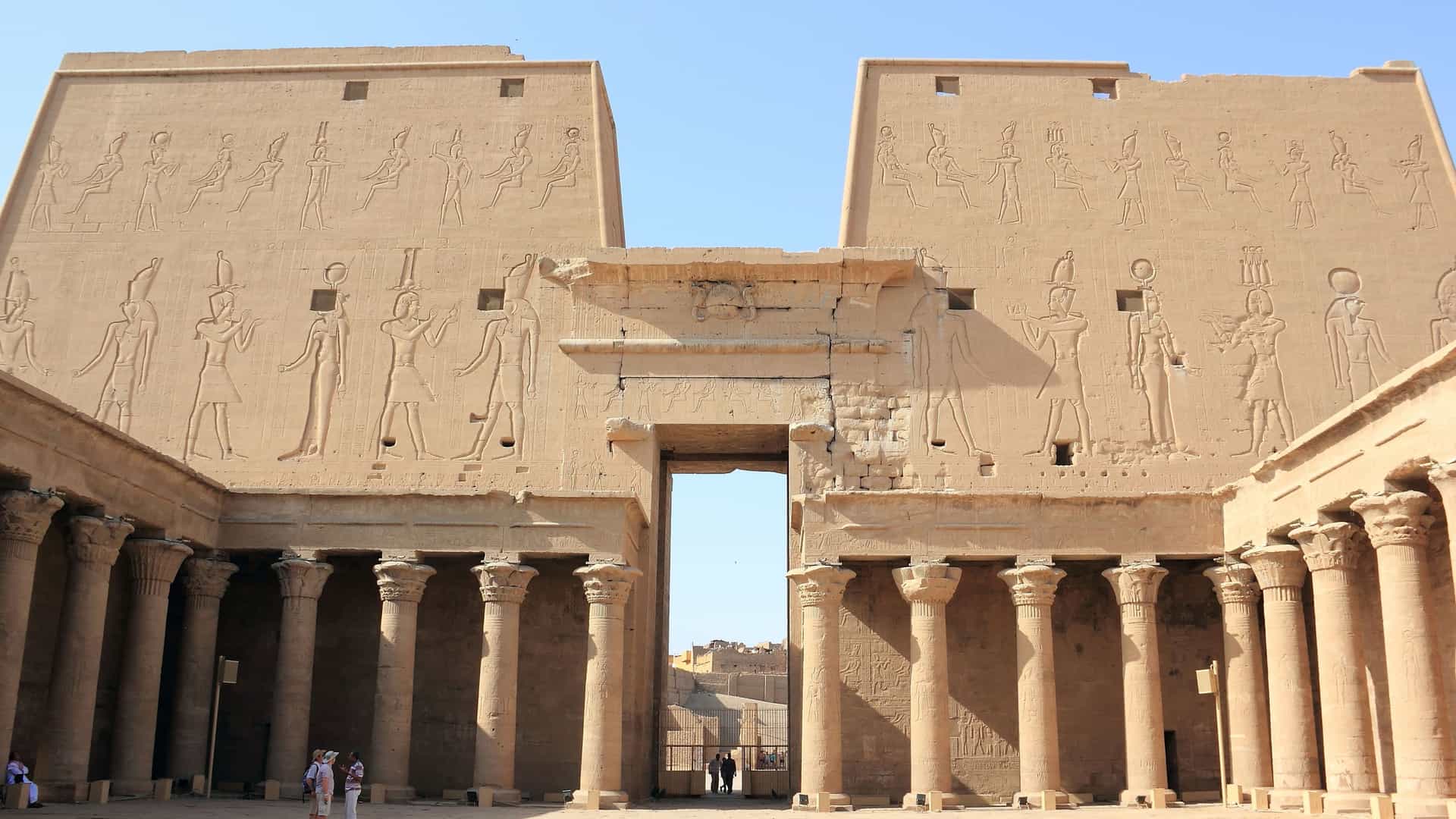 A stunning low-angle view of the ancient Temple of Horus in Edfu, Egypt, with its massive sandstone walls covered in detailed carvings and a colonnaded courtyard.