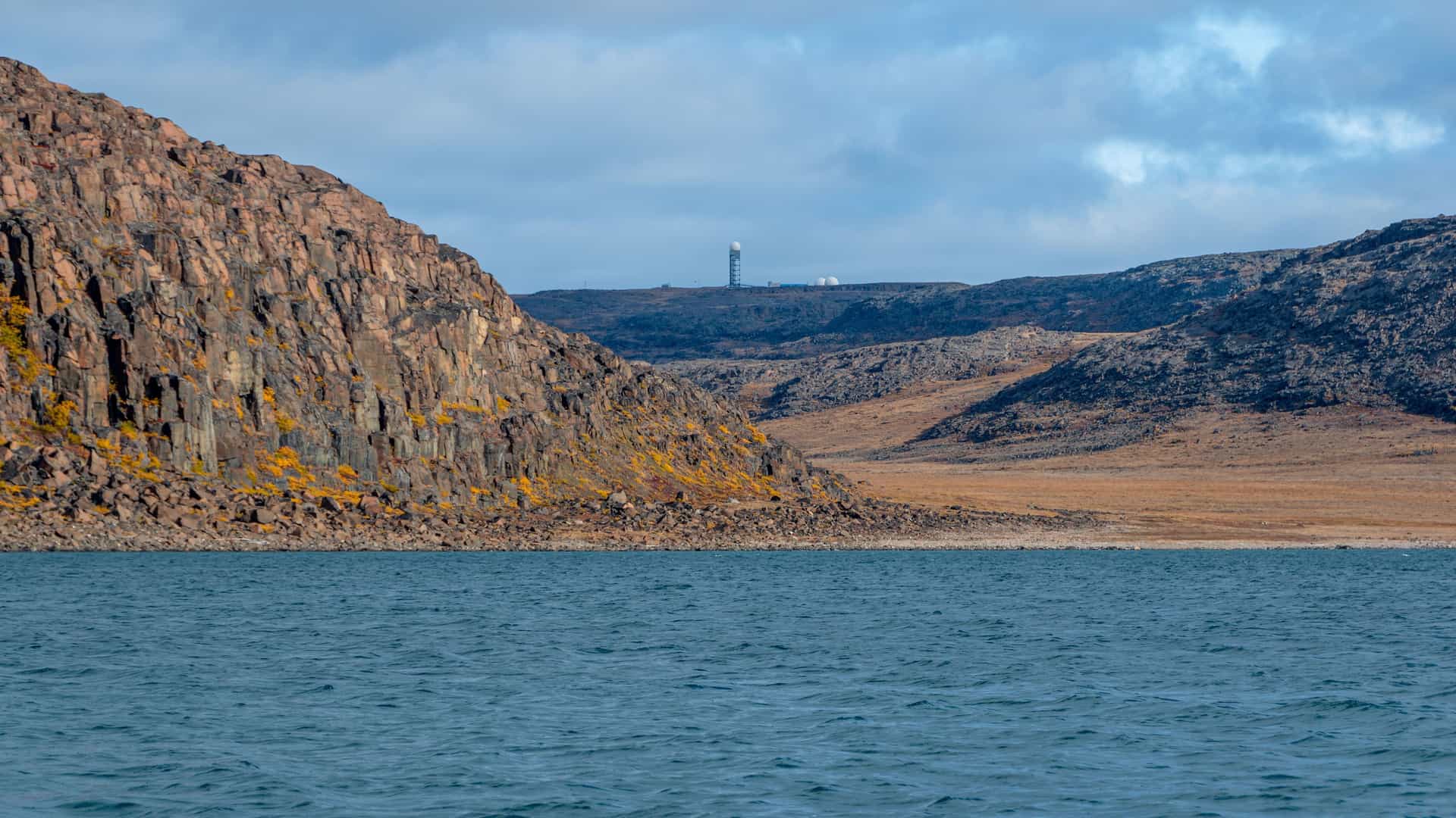 A panoramic shot of the rugged coastline of Edinburgh Island, with rocky cliffs and a barren landscape under a cloudy sky, with a telecommunications tower visible in the distance.