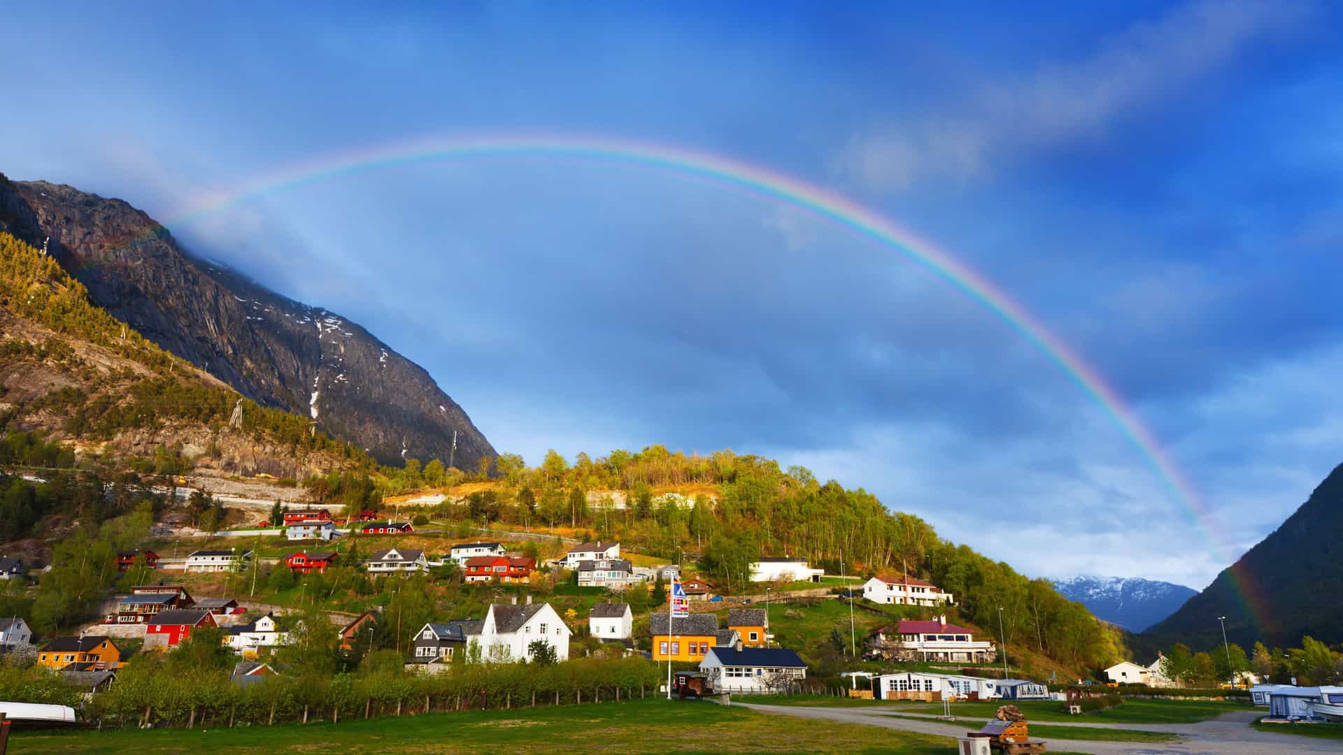 A vibrant rainbow arching over the village of Eidfjord, Norway, with colorful houses nestled on a lush green hillside below a majestic mountain, under a dramatic sky.