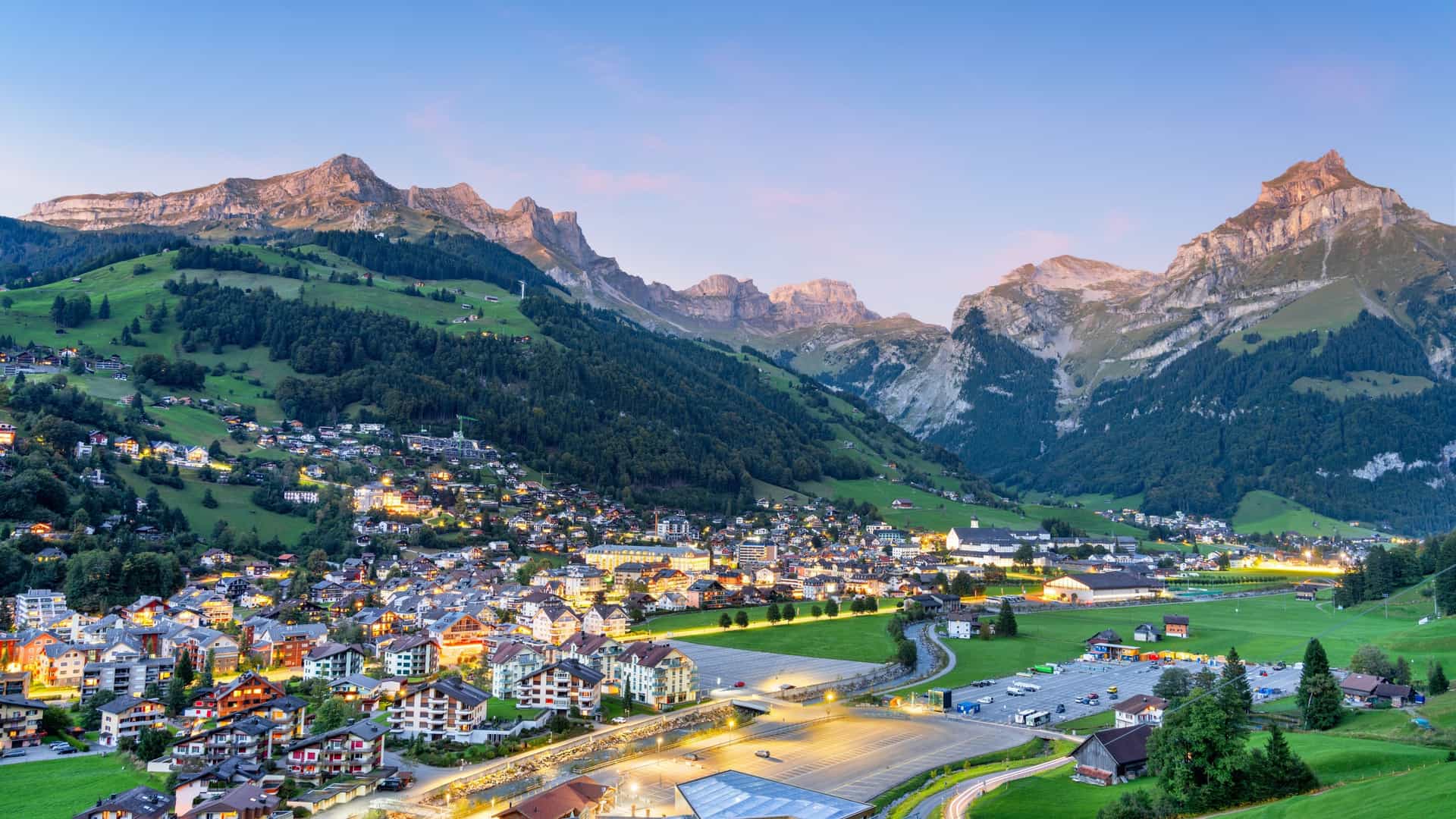 Illuminated alpine village of Engelberg, Switzerland, nestled in the valley beneath the towering peaks of the Uri Alps at dusk.
