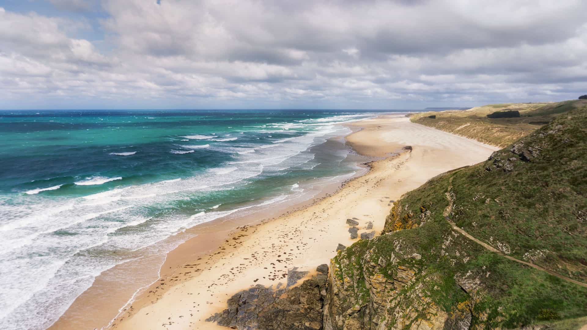 A stunning aerial view of a wide sandy beach on the Bay of Biscay coast, with powerful turquoise waves crashing against the shore and lush green cliffs meeting the sea.
