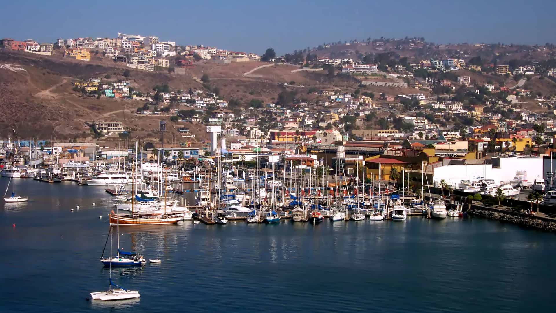 A bustling marina in Ensenada, Mexico, filled with sailboats and yachts, with the colorful buildings of the city climbing up a barren hillside in the background.