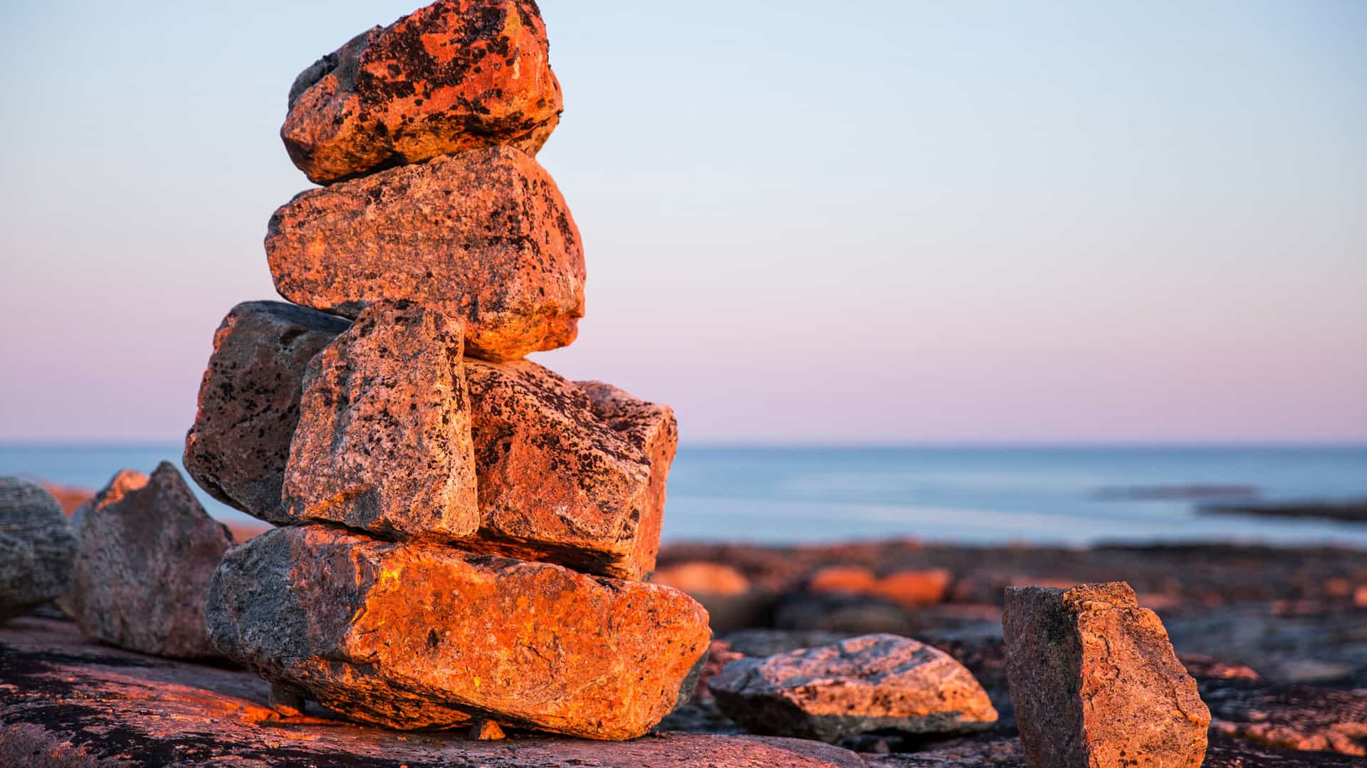 "A low-angle shot of a cairn, a stack of large, reddish-colored rocks, on the shore of Erik Harbour, Nunavut, with the calm blue ocean and a clear sky with a hint of pink at the horizon in the background.  "