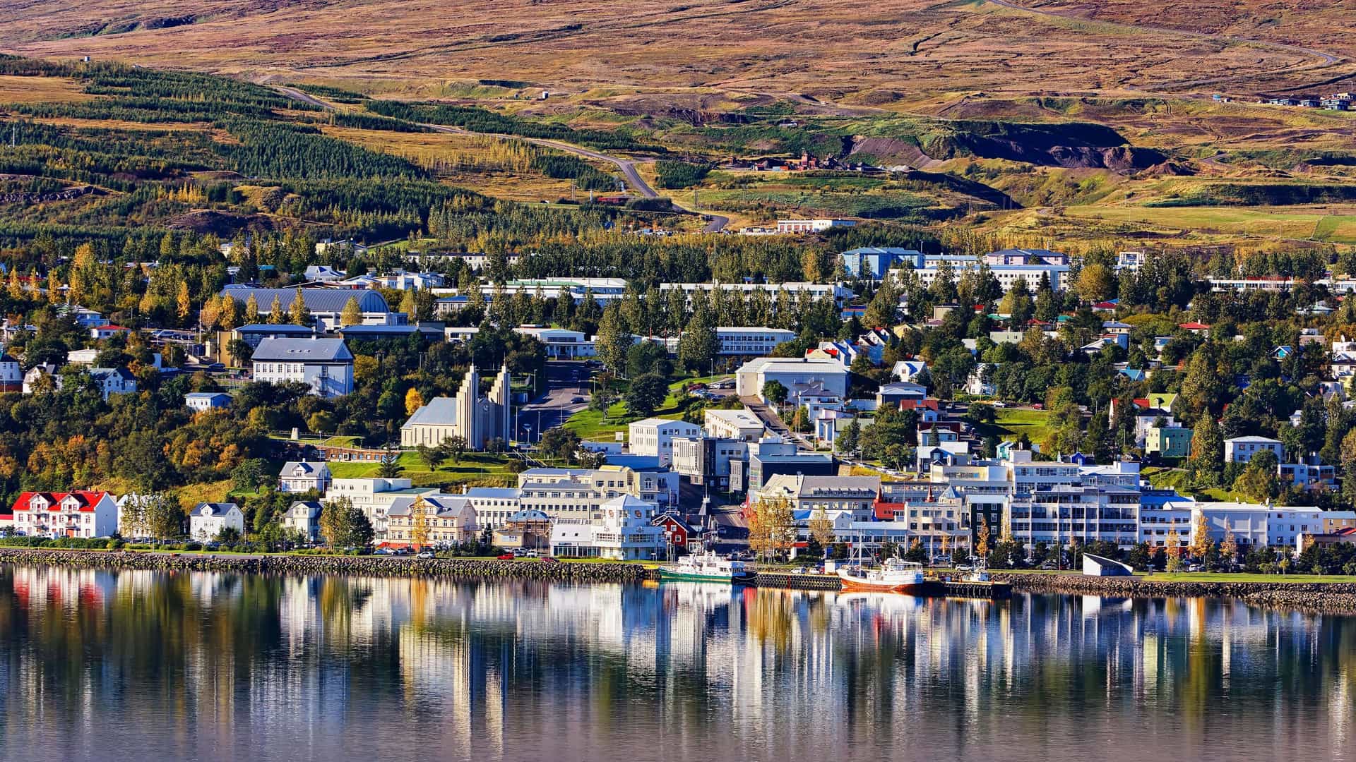 A scenic view of Akureyri, a town in northern Iceland, nestled between rolling hills and the tranquil waters of Eyjafjörður, with buildings and mountains beautifully reflected on the water's surface.
