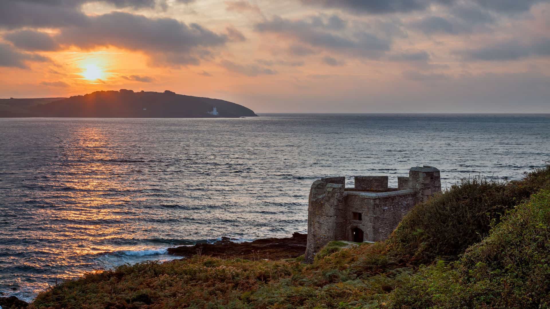 A breathtaking sunset over the sea near Falmouth, Cornwall, with a historical stone fort in the foreground and a distant coastline silhouetted against the bright, cloudy sky.