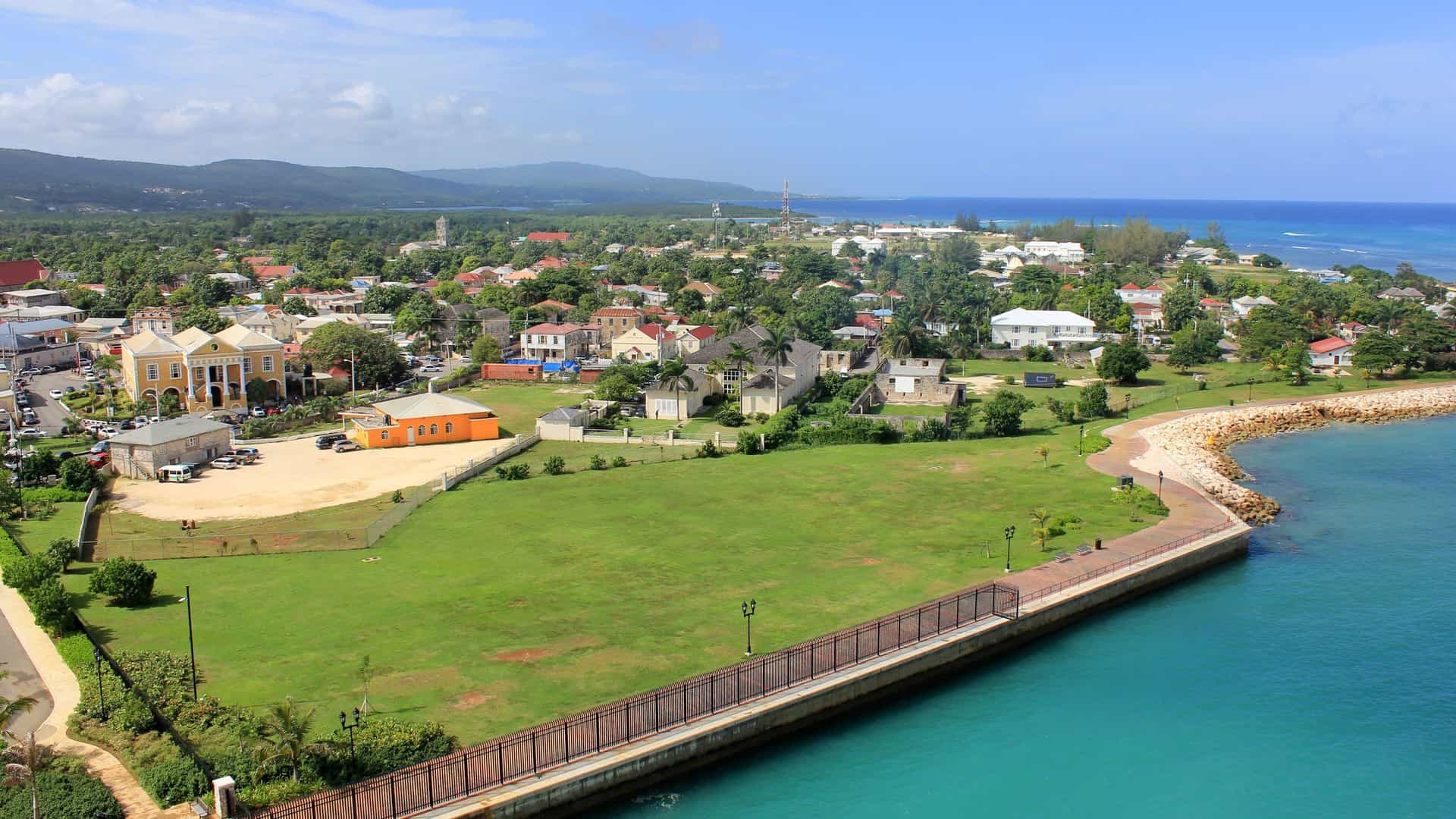 An aerial view of the historic town of Falmouth, Jamaica, showing a coastal promenade along the turquoise sea, colorful colonial-era buildings, and lush green hills in the background.