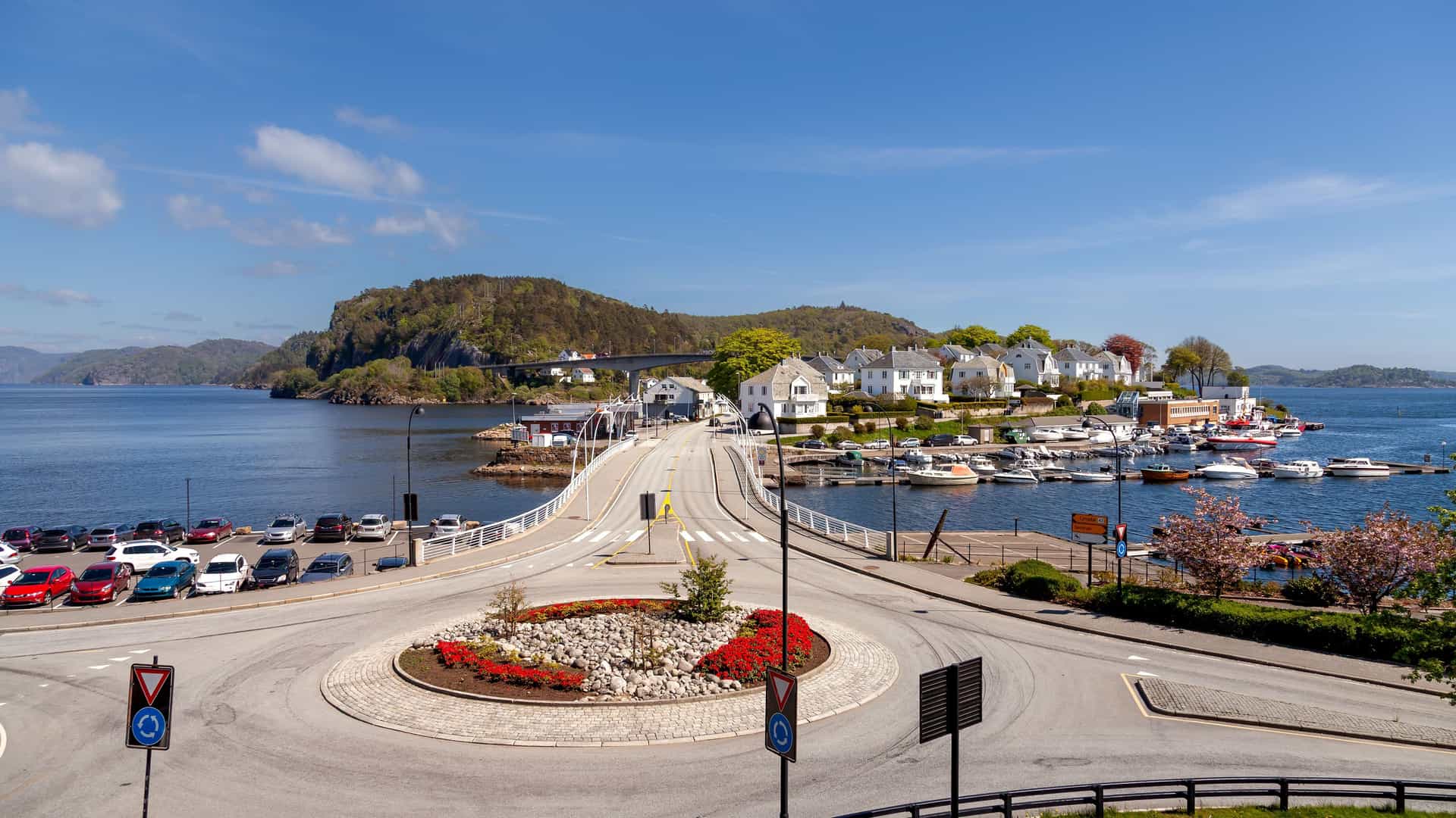 "A sunny panoramic shot of Farsund, Norway, with a busy roundabout in the foreground leading to a bridge connecting the mainland to a small island filled with white houses, a bustling marina, and lush green hills.  "
