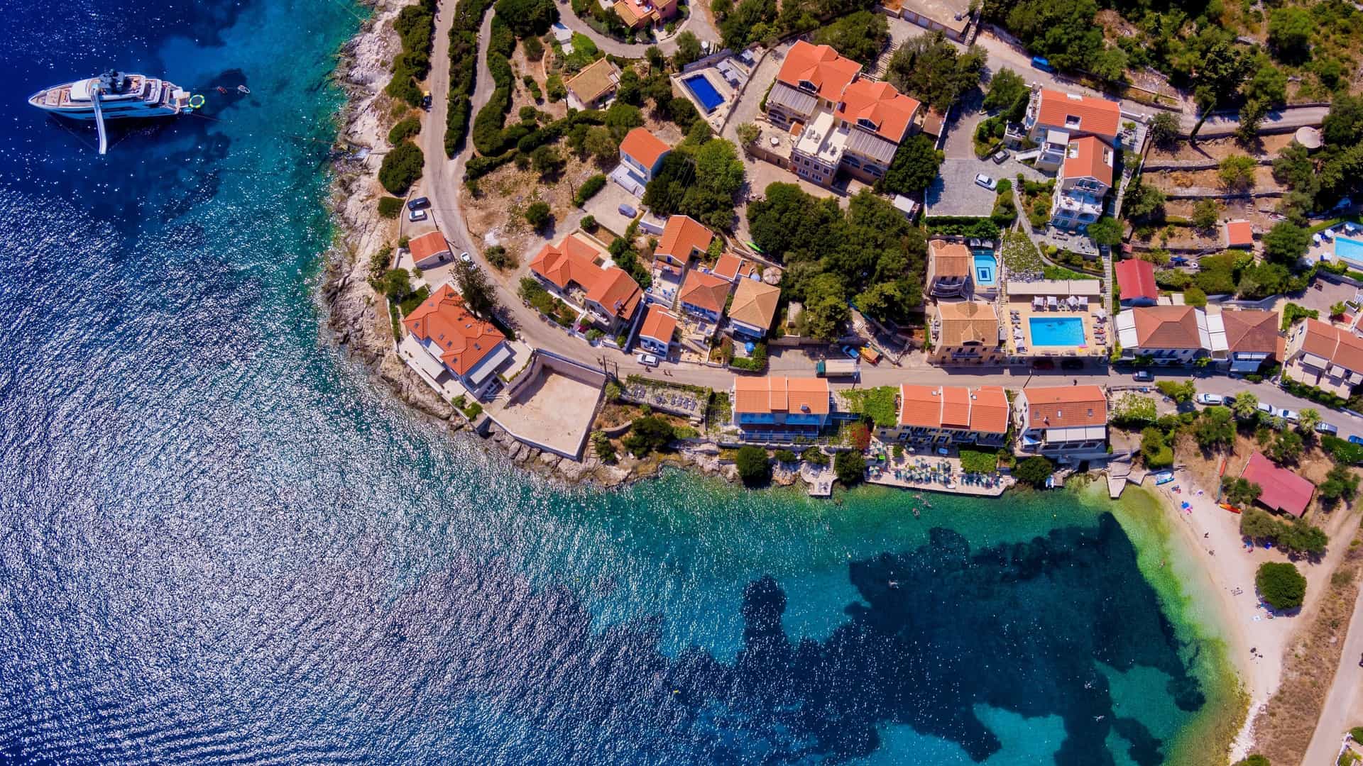 An aerial view of the picturesque Fiskardo village and port on Kefalonia island, Greece. Colorful houses with red tiled roofs line the crescent-shaped harbor, where several yachts and sail boats are moored in the bright turquoise-blue water. The village is nestled on a peninsula surrounded by lush green hills.