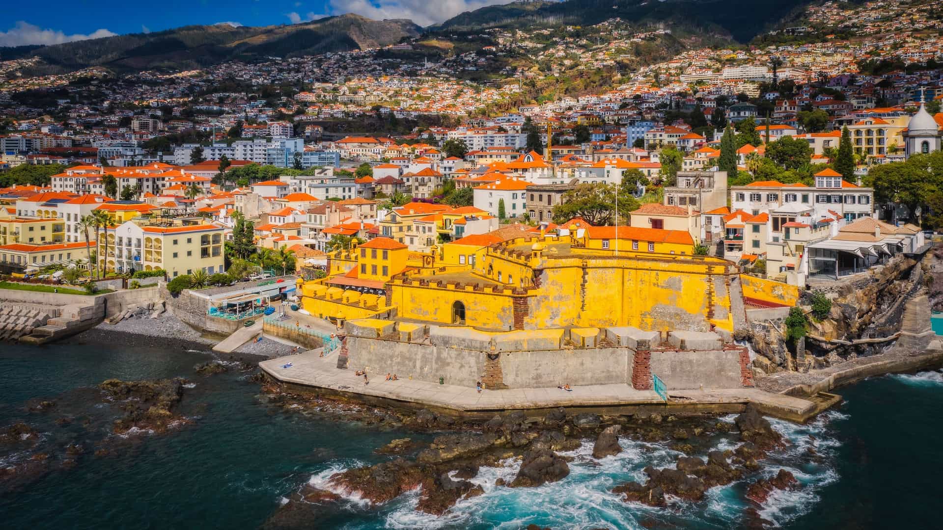 An aerial view of Fortaleza de São Tiago, a historic yellow fortress, on the coastline of Funchal, Madeira, with the densely packed city and green mountains in the background.