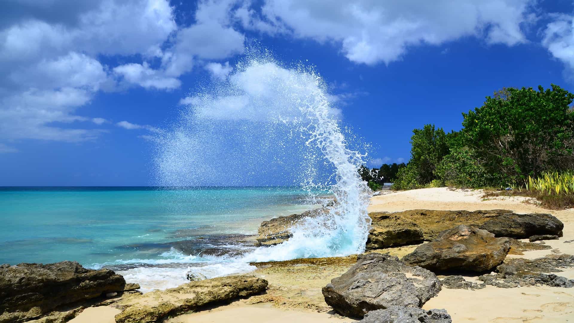 A vibrant tropical scene on the beach of Frederiksted, St. Croix, with a wave crashing spectacularly against the rocky shoreline under a bright blue sky with white clouds.