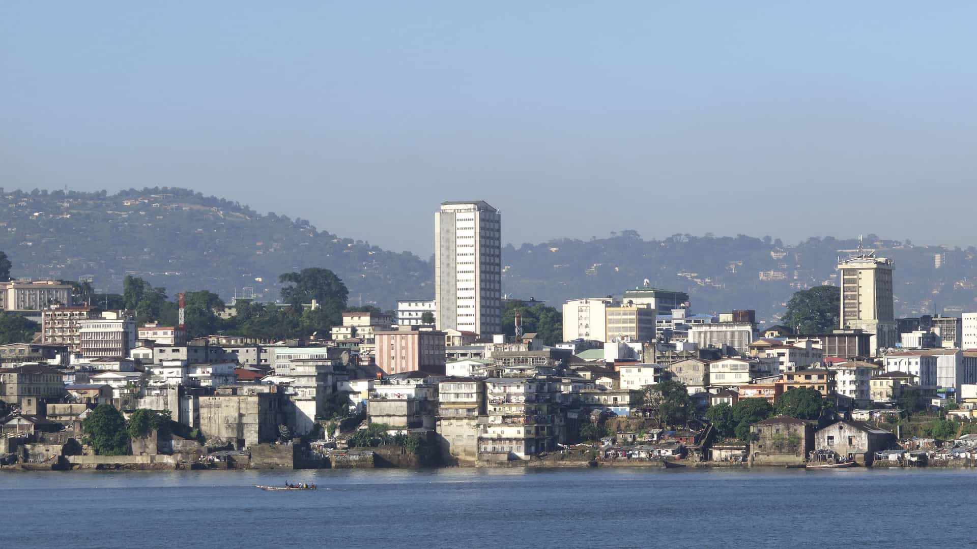 A view of the Freetown, Sierra Leone skyline from the water, showing a mix of modern high-rise buildings and older waterfront structures against a backdrop of green hills.