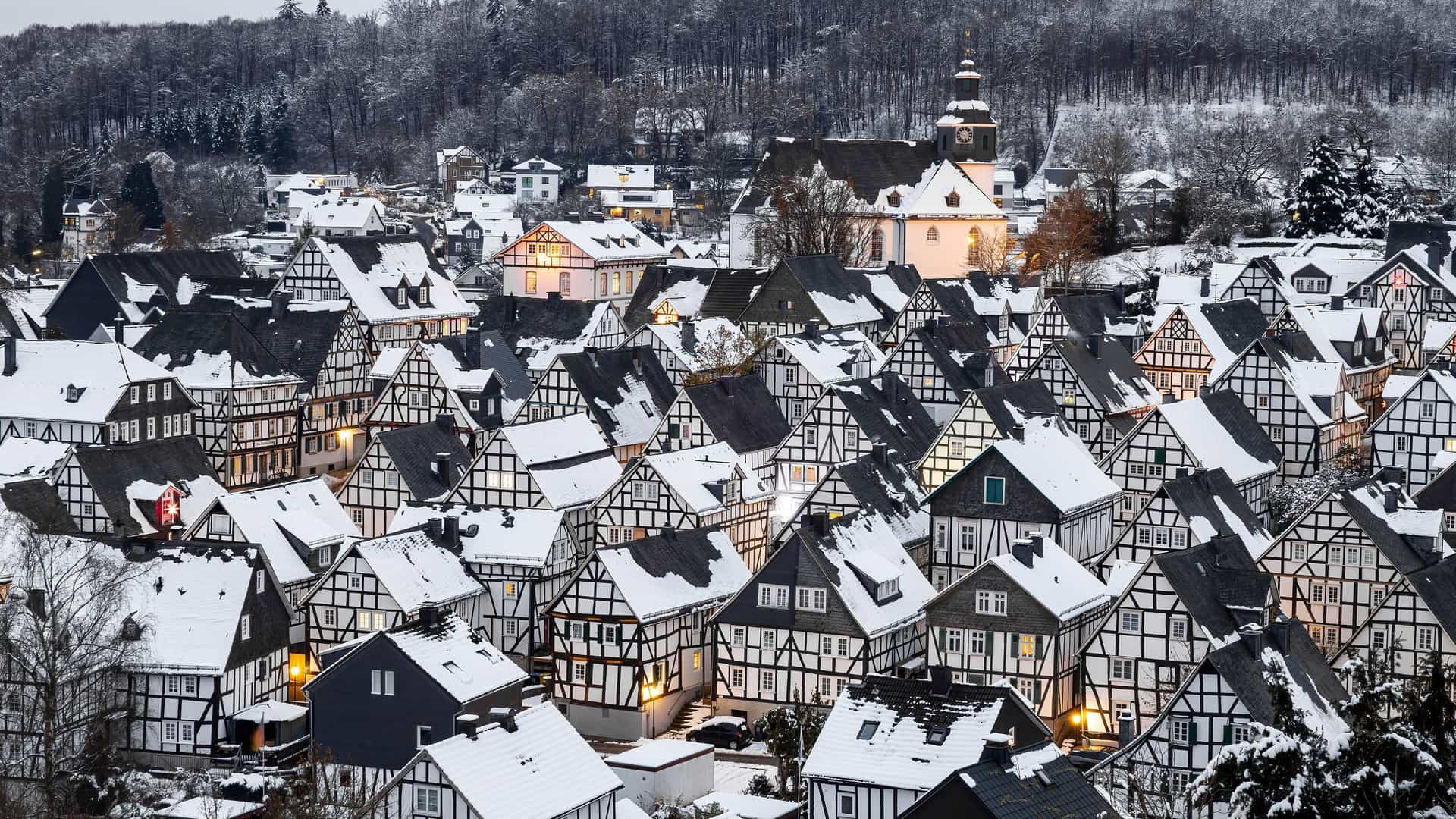 A wide-angle, elevated shot of the old town of Freudenberg, Germany, with its iconic black-and-white half-timbered houses blanketed in snow under a winter sky.