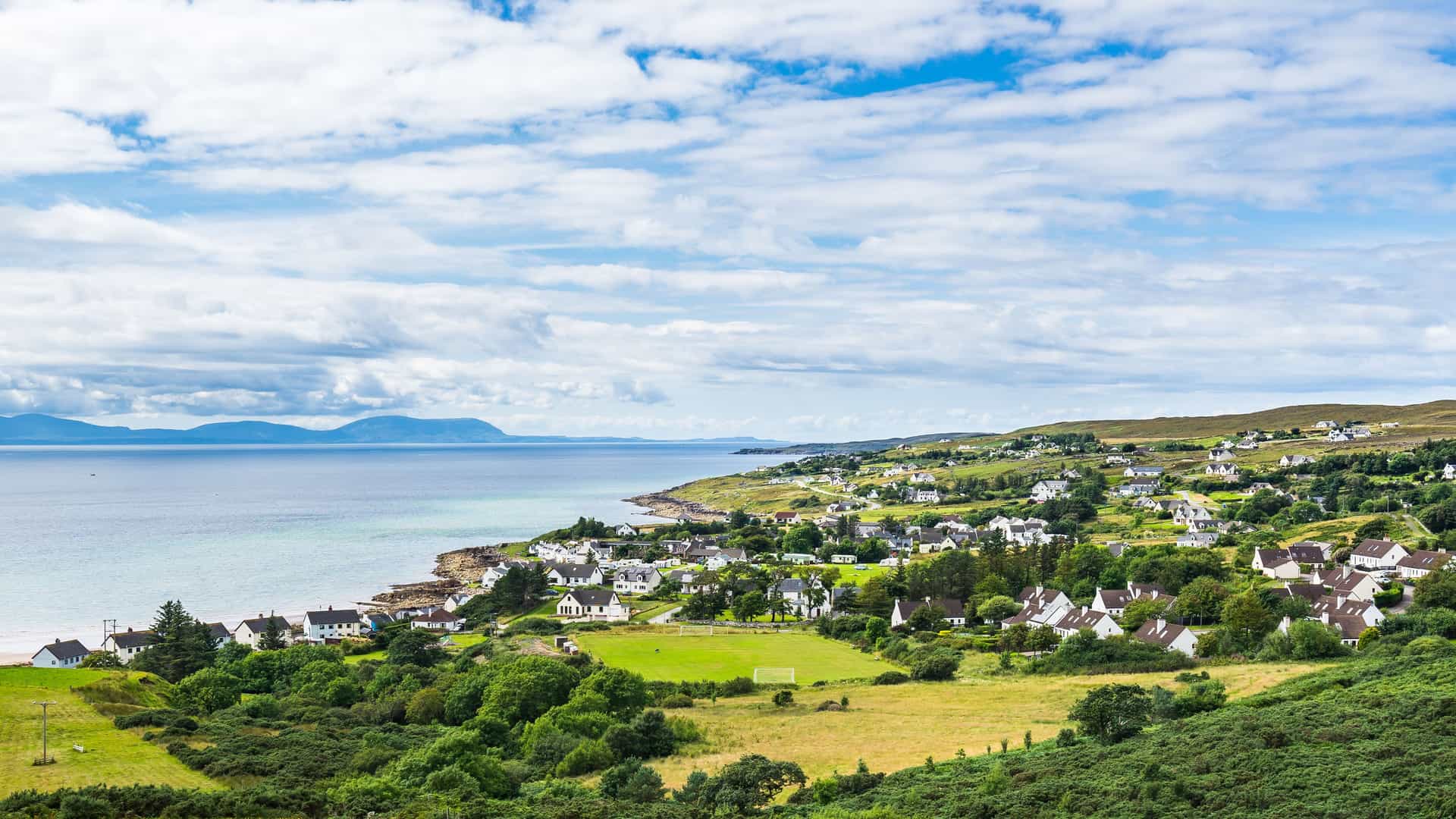 "A beautiful panoramic shot of the coastal village of Gairloch in Scotland, with white houses scattered across green hills overlooking the serene loch and distant mountains. "