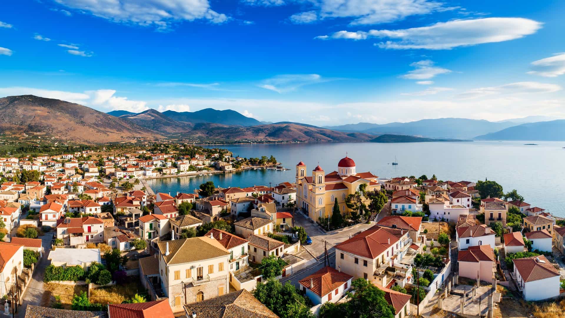  A stunning wide-angle view of the seaside town of Galaxidi, Greece, featuring houses with red-tiled roofs and a church with a distinct red dome, set against the blue Gulf of Corinth and mountains.