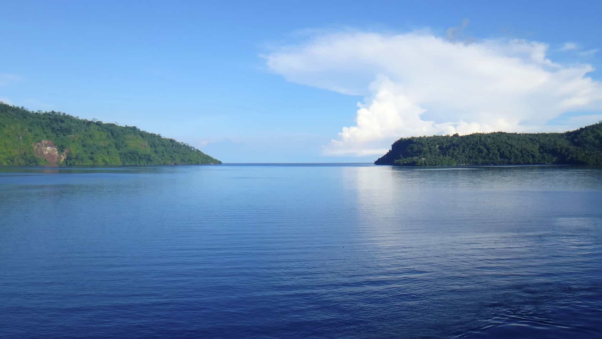 A tranquil scenic shot of a calm blue ocean channel with two lush, green islands on either side under a beautiful blue sky with a few clouds.