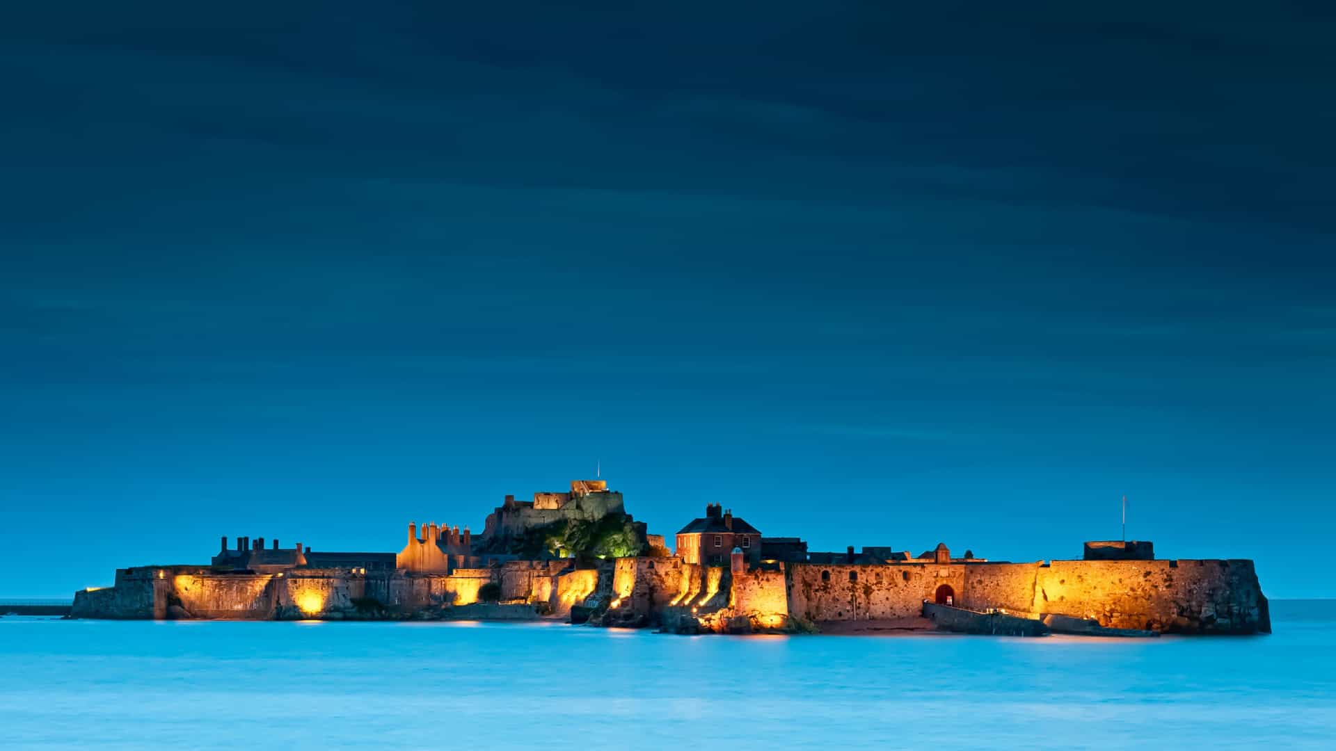 A historic castle fortress illuminated at night, located on a small island surrounded by calm water under a deep blue evening sky.