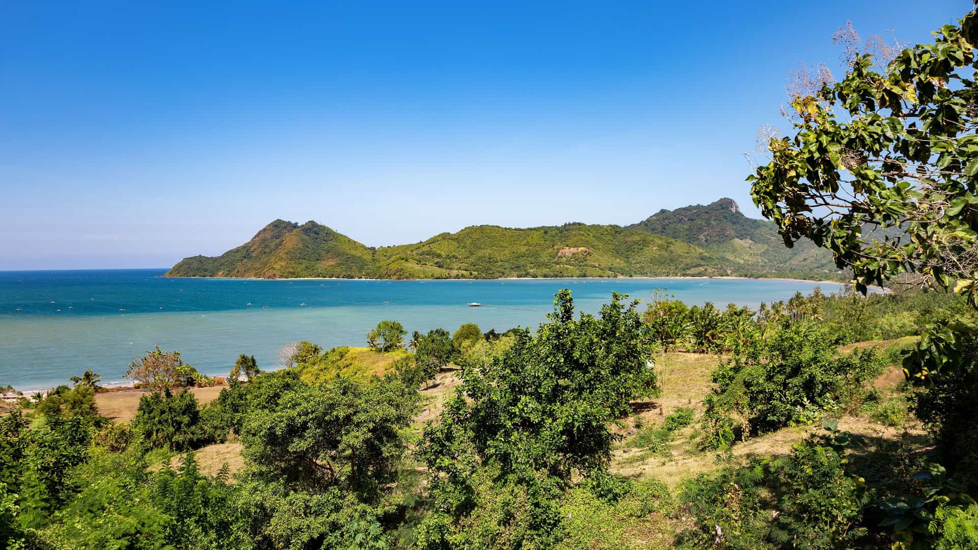 A panoramic view of Gili Mas Island, Indonesia, with a turquoise bay, lush green mountains, and a beautiful sandy beach on a sunny day.